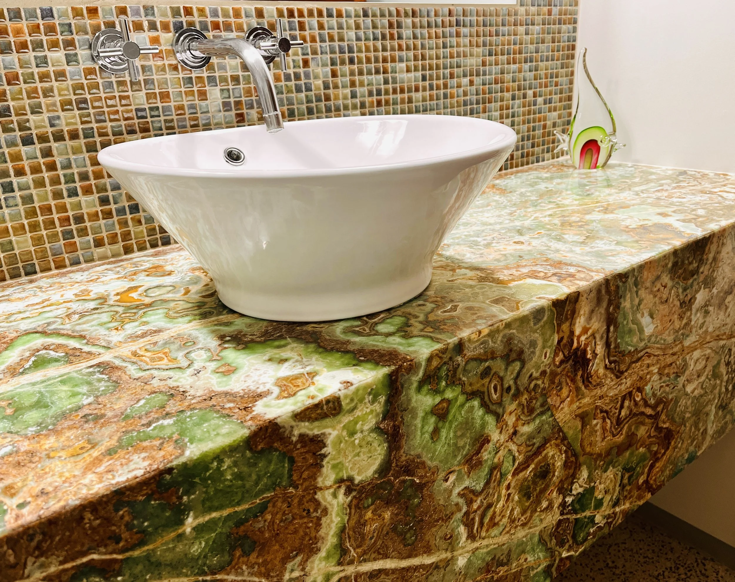 Bathroom sink with a white vessel basin, chrome faucet, and colorful mosaic tile backsplash on a granite countertop with green and brown patterns. A decorative glass vase is placed on the right.