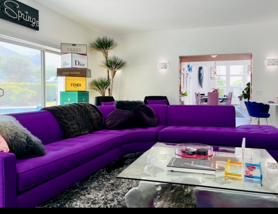 Bright living room with purple sectional sofa, black and fuzzy pillows, glass coffee table with remote controls, magazines, and colorful markers, window with a view of trees, and trees on a shelf near the window.