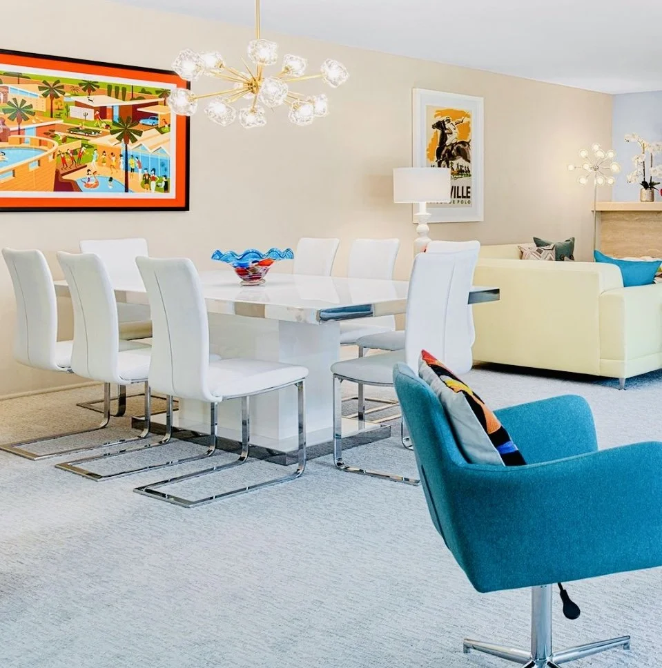 Modern dining room with white dining table surrounded by eight white chairs, a colorful wall art, a gold chandelier, a yellow sofa, a blue armchair, and modern table lamps.