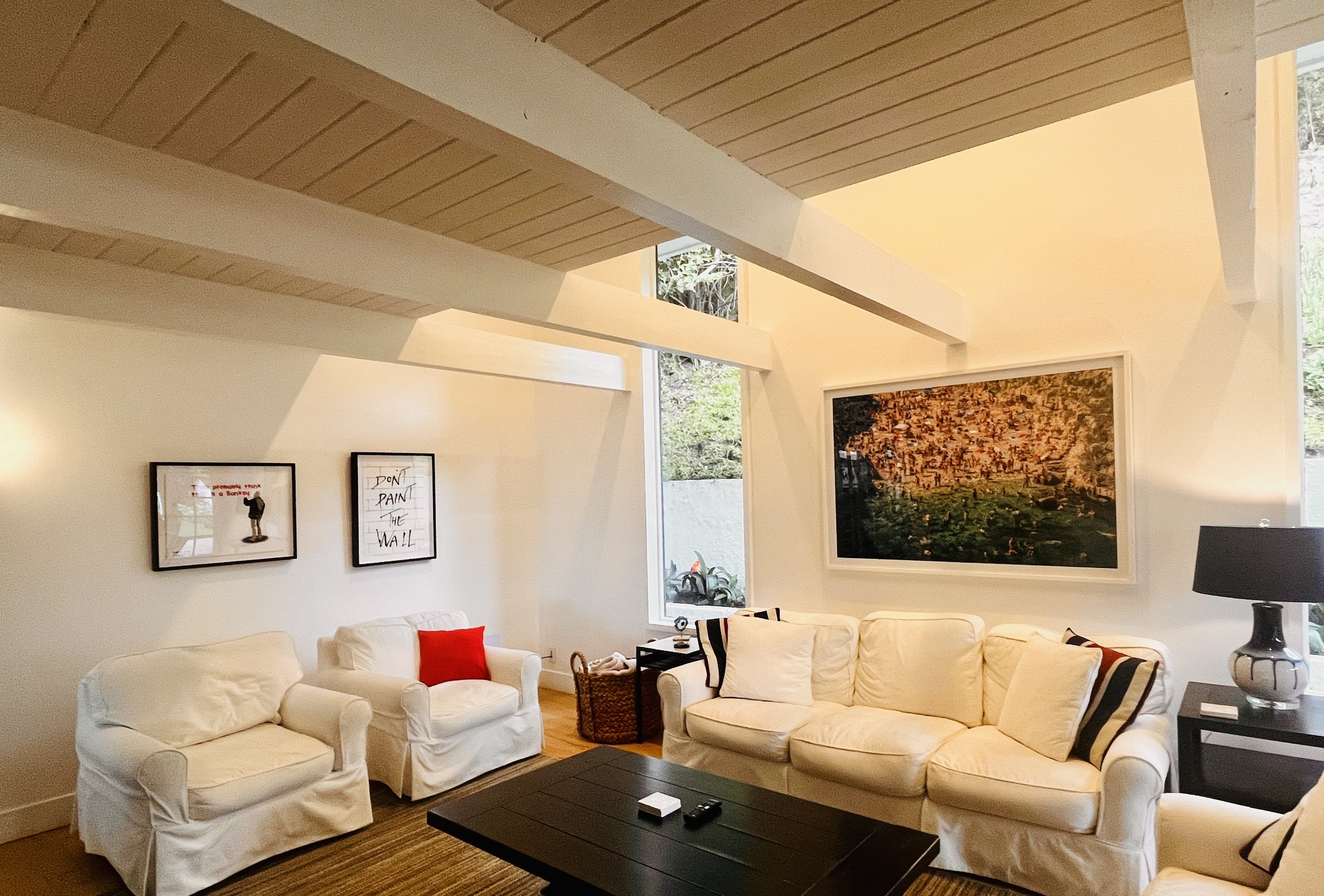 Living room with white sofas, a black coffee table, wall art including a large framed cityscape painting, and a window showing trees outside.