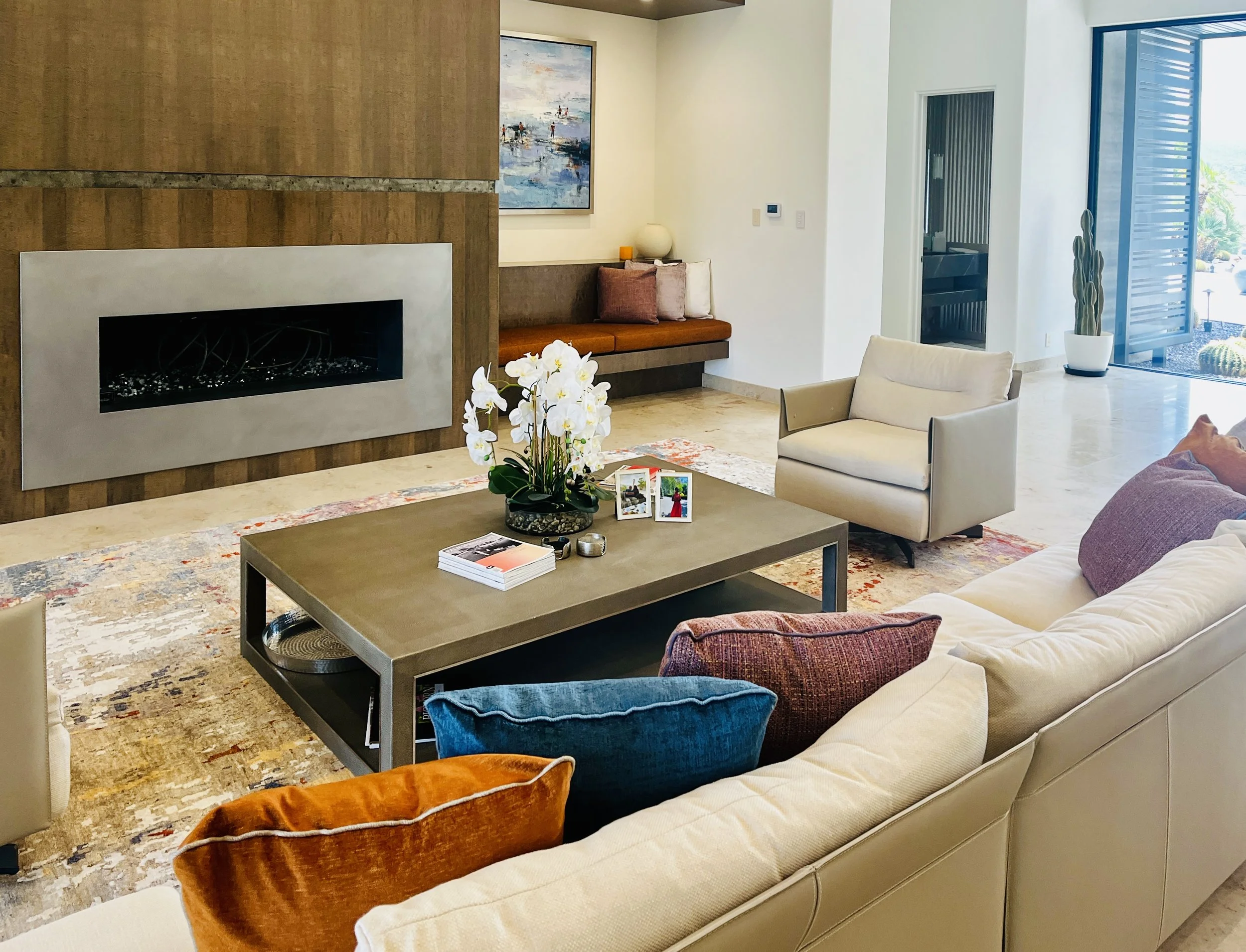 Living room with beige sofa, colorful cushions, coffee table, indoor plants, fireplace, artwork, and seating area near large glass door.