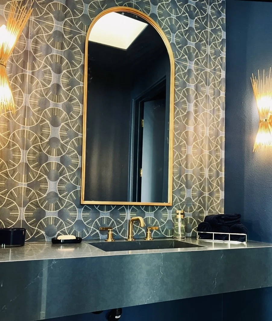 A modern bathroom vanity with a dark green marble countertop, a gold faucet, and a decorative gold-framed mirror. The wall behind features patterned wallpaper with circular geometric designs in dark blue and gray tones, flanked by gold wall sconces w
