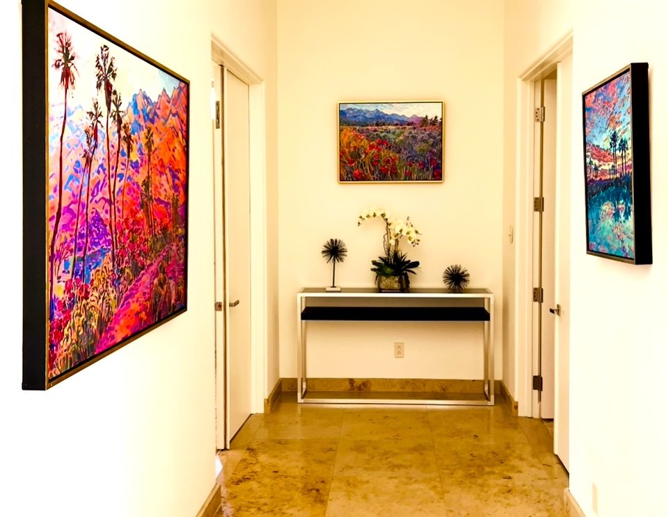 Hallway with three colorful landscape paintings on cream walls and a white table with plants.