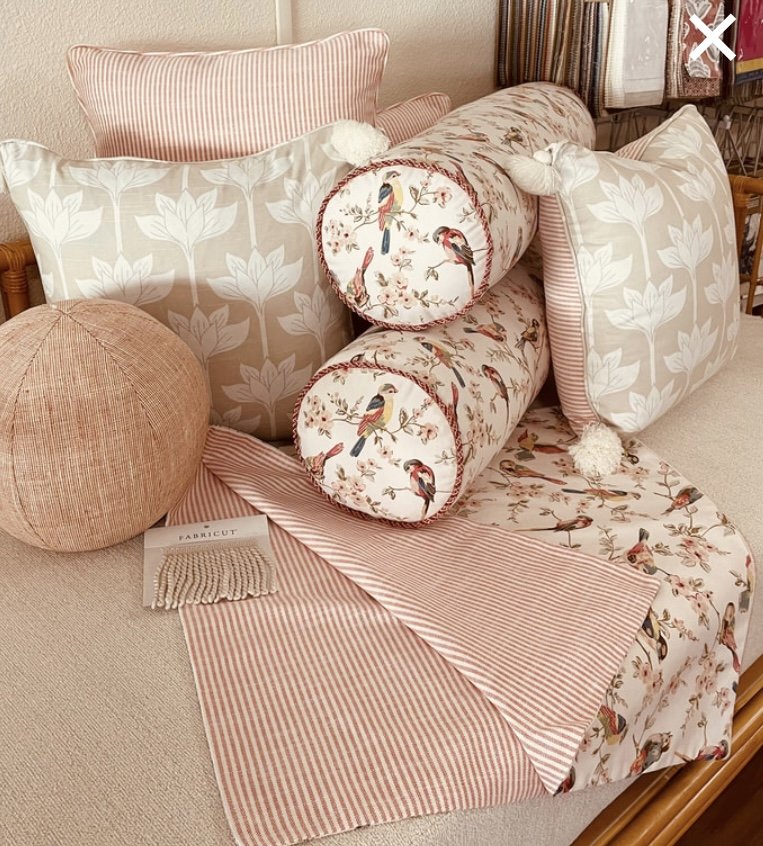 Arrangement of decorative pillows, a round fabric ball, and a fabric sample on a beige upholstered couch. The pillows feature floral and bird patterns, with some striped and leaf designs in soft pink, beige, and cream colors.