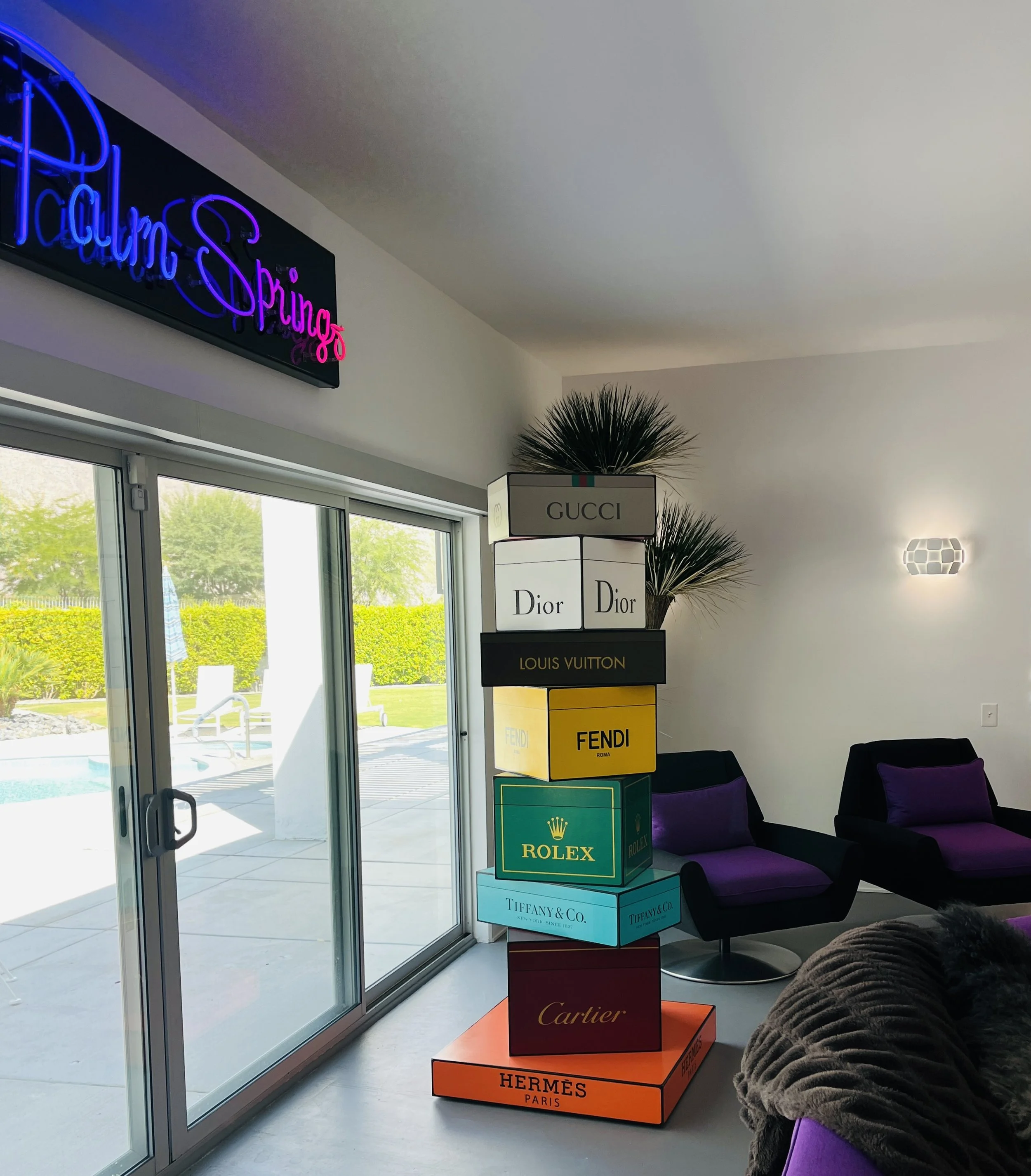 Stacked luxury brand boxes including Gucci, Dior, Louis Vuitton, Fendi, Rolex, Tiffany & Co., Cartier, and Hermès, near sliding glass doors leading to an outdoor pool area.