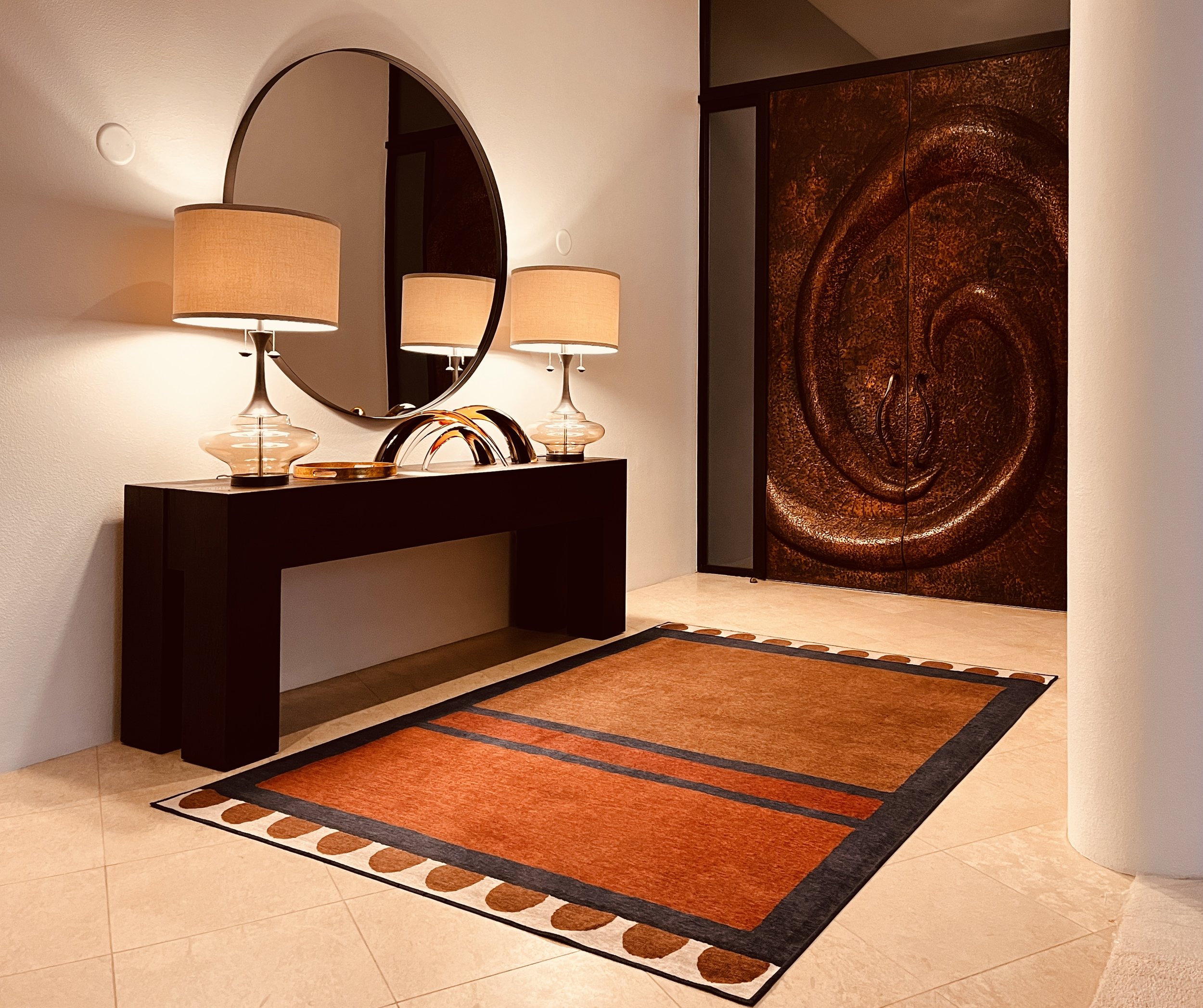 Interior of a hotel or modern home entryway with a black console table, two table lamps with beige shades, a round mirror, a decorative sculpture, a patterned area rug, and a large wooden door with a swirling pattern.
