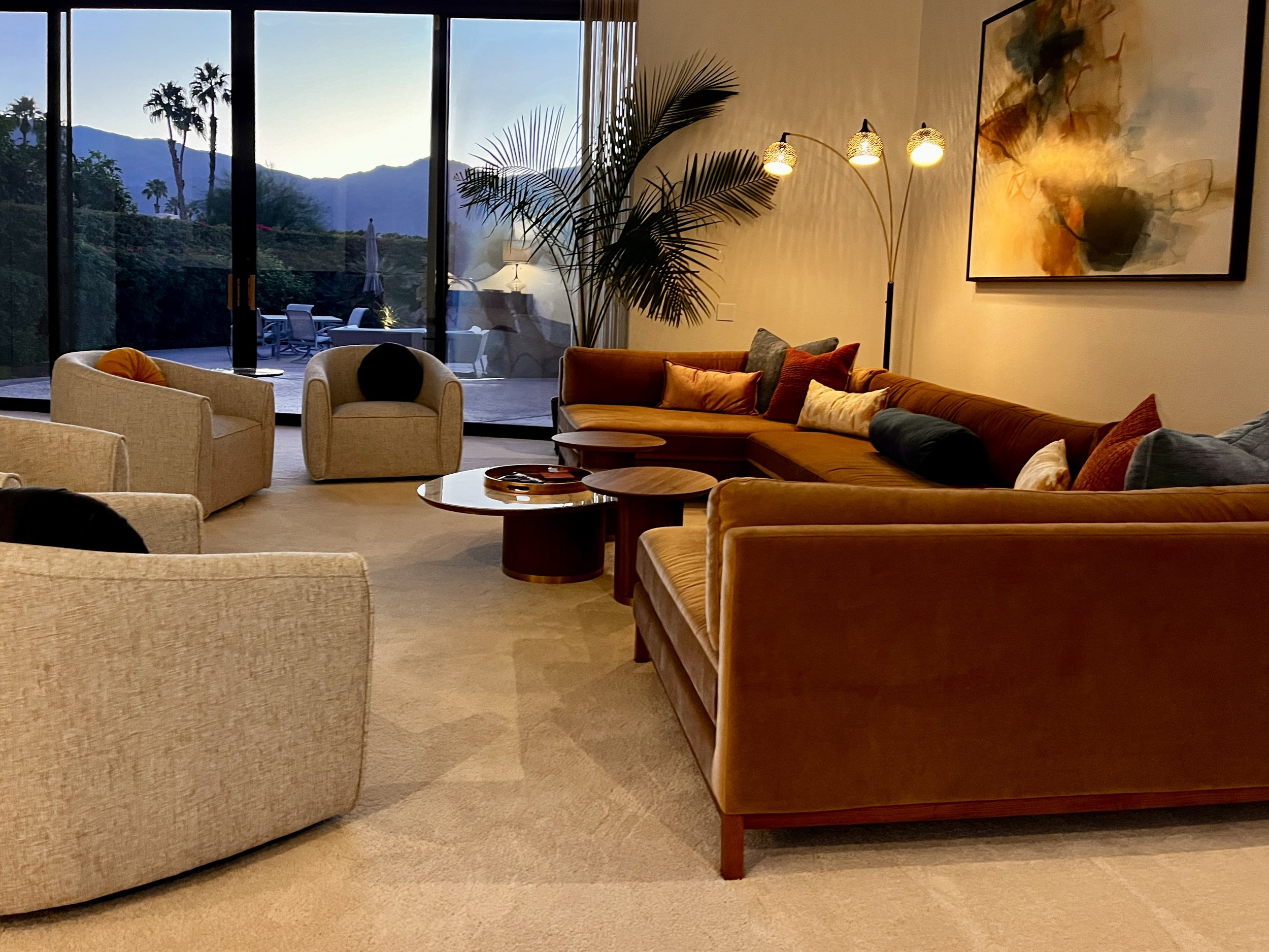 Living room with beige and brown sofas, a round coffee table, a floor lamp, artwork on the wall, large glass sliding doors showing an outdoor patio with mountains and palm trees at sunset.