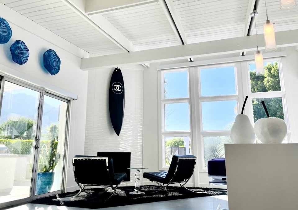 Modern living room with large windows, black chairs around a small glass table, a surfboard with Chanel logo on the wall, blue wall art, and decorative white vases with black straws.