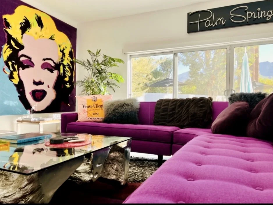 Living room with a bright pink sectional sofa, black and furry throw pillows, a glass coffee table with various items on it, a large colorful pop art portrait on the wall, a potted plant, and a window showing trees and mountains outside. There's a bl