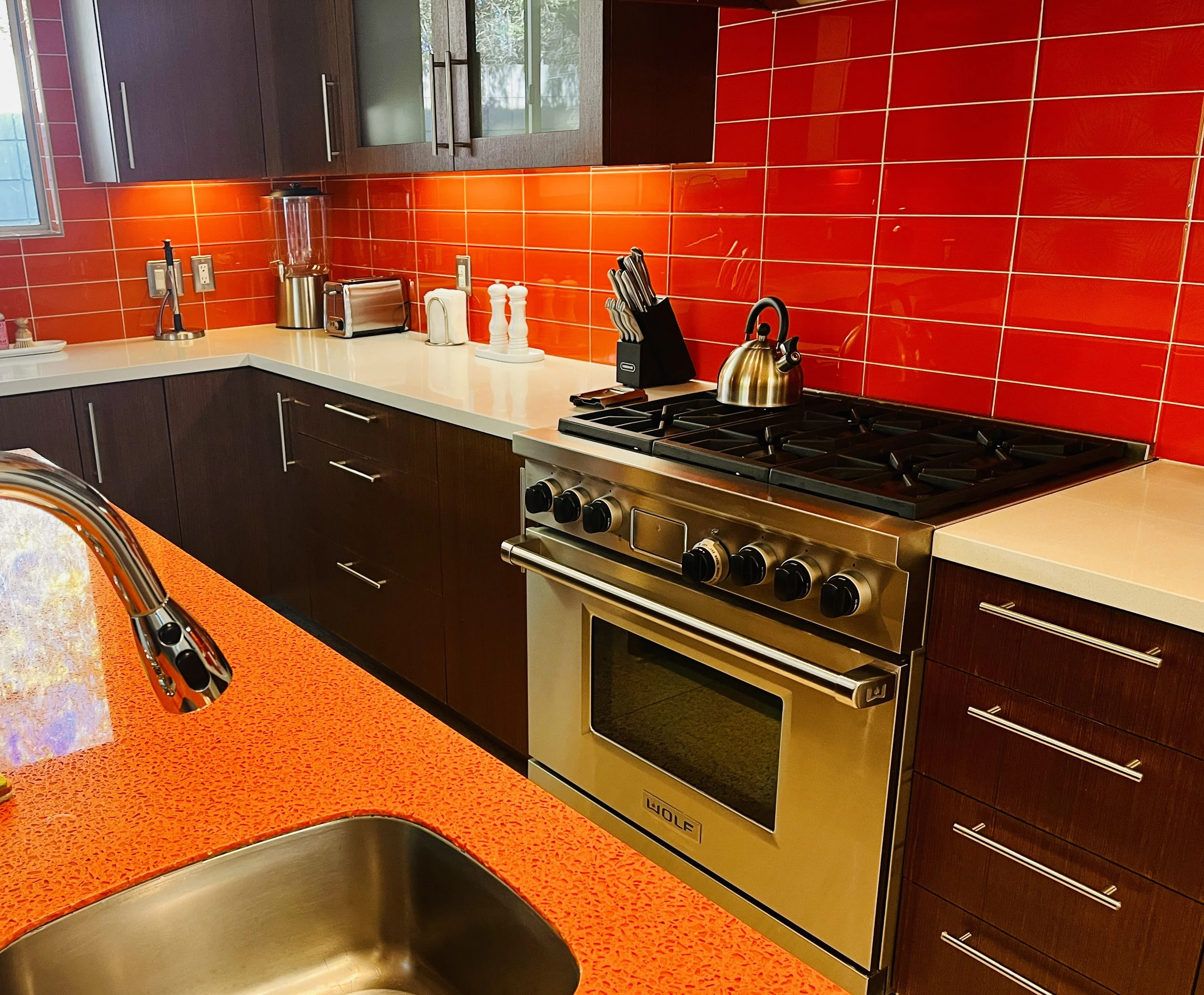 Modern kitchen with red tile backsplash, dark wood cabinets, stainless steel appliances, and an orange speckled countertop with a stainless steel sink.