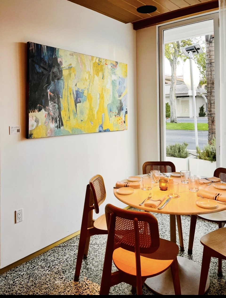 A cozy dining area with a round wooden table set with plates, glasses, and napkins, surrounded by wooden chairs. A large abstract painting hangs on a white wall, and there is a large window with a view of a street and trees outside.