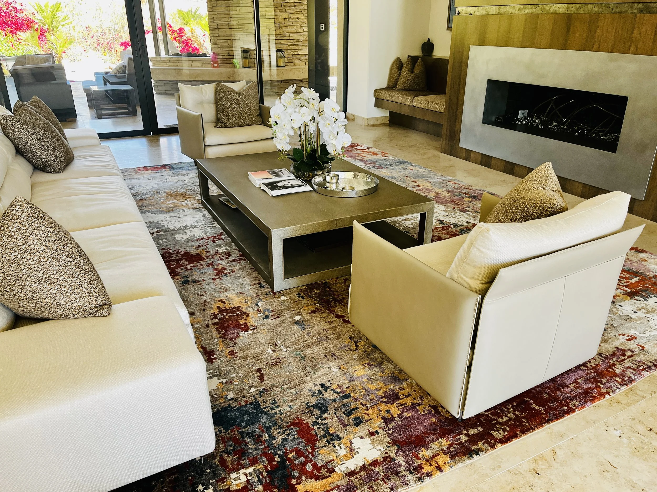 Living room with cream-colored sofa and armchairs, patterned pillows, a metal coffee table with books and a white orchid, a colorful area rug, and a fireplace with a wooden surround.