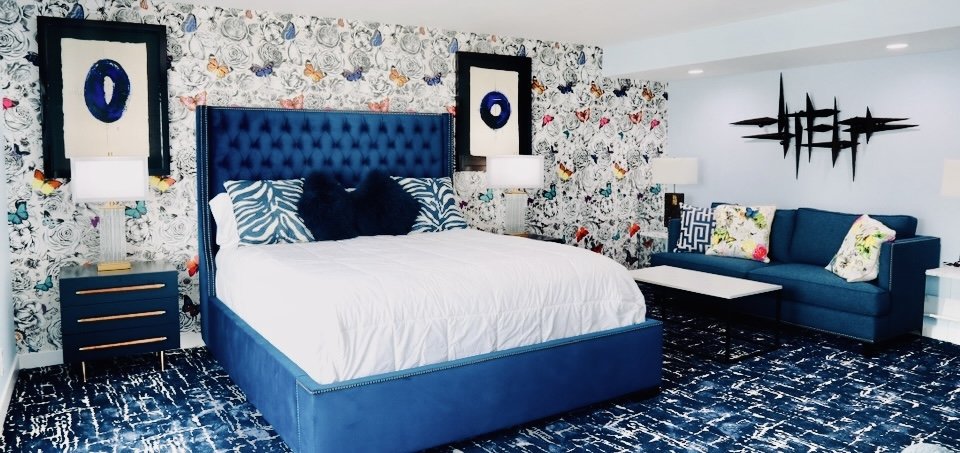 A bedroom with a blue bed, floral and butterfly wallpaper, a navy blue sofa with colorful pillows, and wall art.