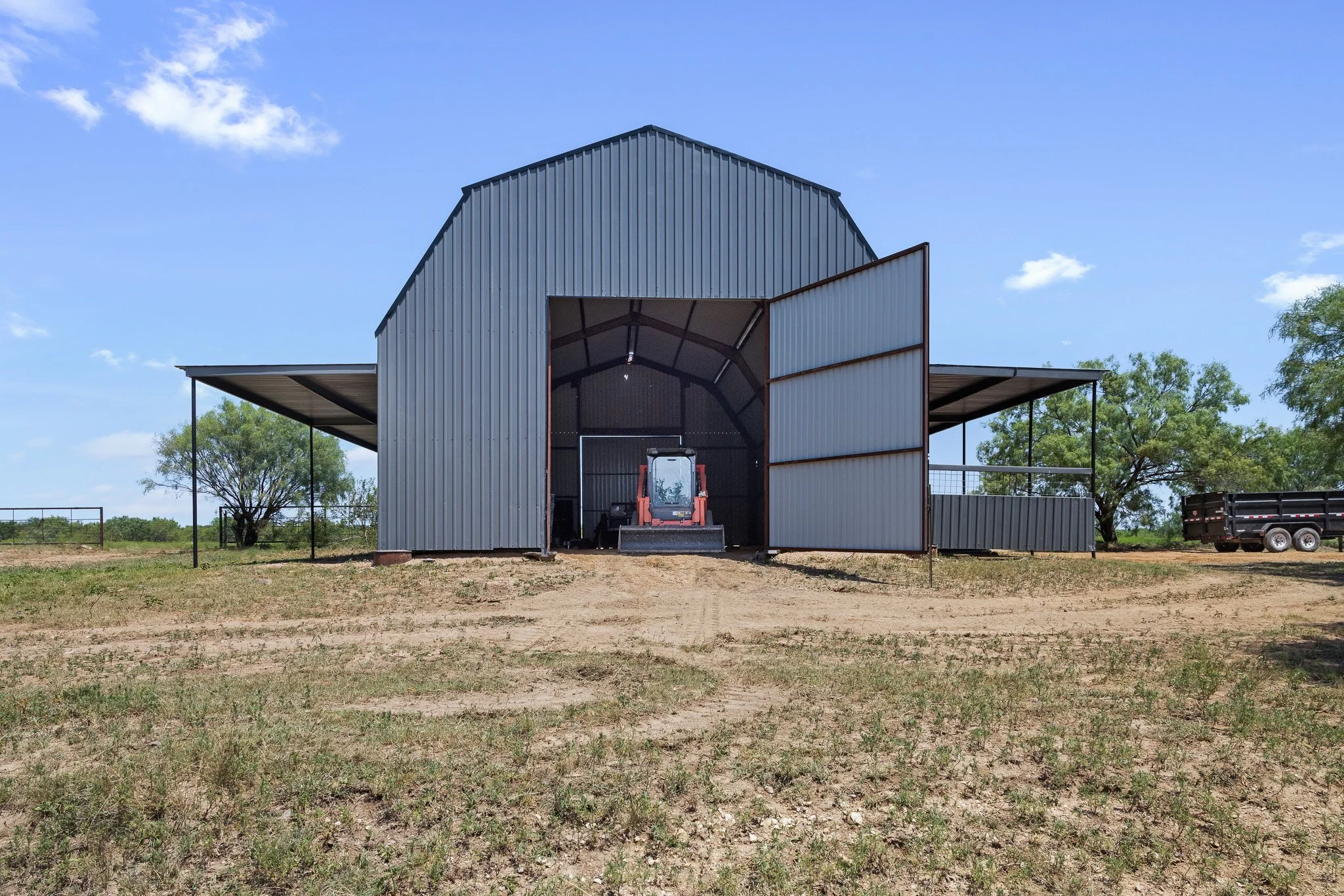 Skycrafted Imaging_Real Estate Media Production_Central & Southeast Texas_A metal shed with large open doors revealing a front loader inside, on a farm with trees and open land under a partly cloudy blue sky.