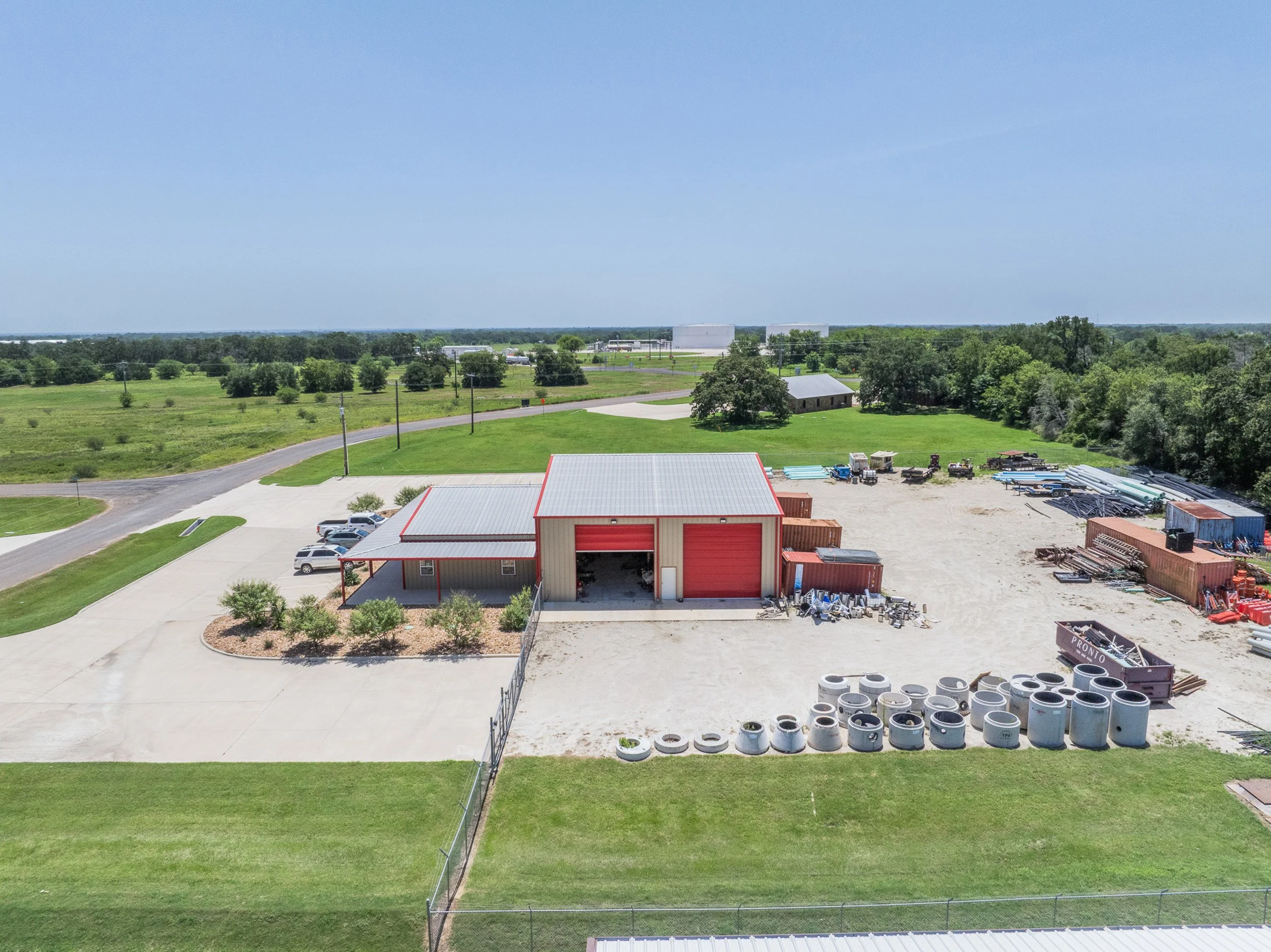 Skycrafted Imaging_Real Estate Media Production_Central & Southeast Texas_Aerial view of a commercial area with a red and beige storage building, vehicles, and pipes outdoors, surrounded by green trees and fields under a blue sky.
