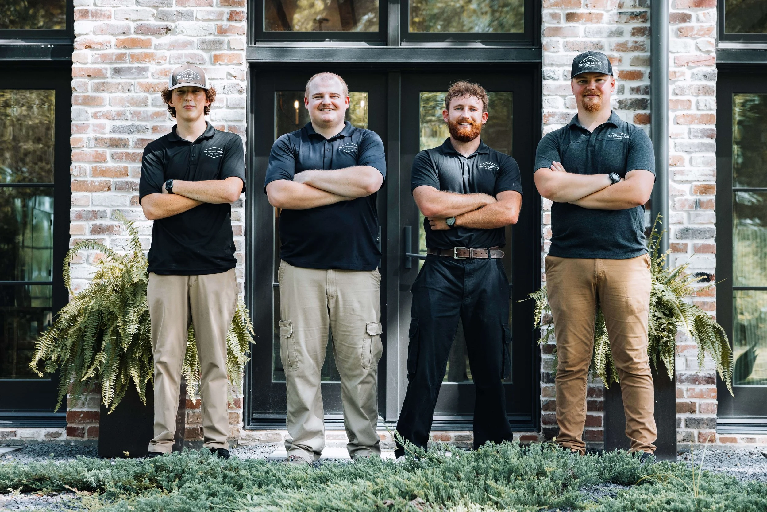 Skycrafted Imaging_Real Estate Media Production_Central & Southeast Texas_Four men standing with arms crossed in front of a brick building with large windows, surrounded by greenery.