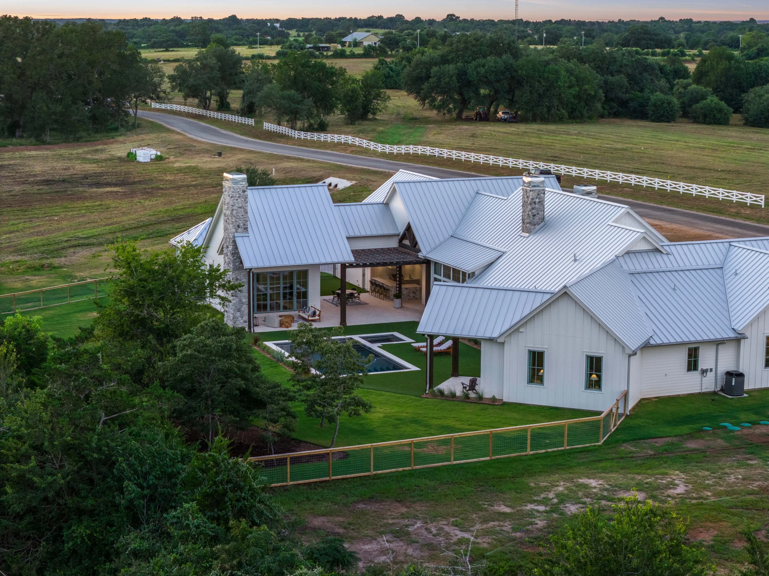 Skycrafted Imaging_Real Estate Media Production_Central & Southeast Texas_A large white house with a gray metal roof, chimney, and outdoor patio, situated on a grassy backyard enclosed by a wooden fence.