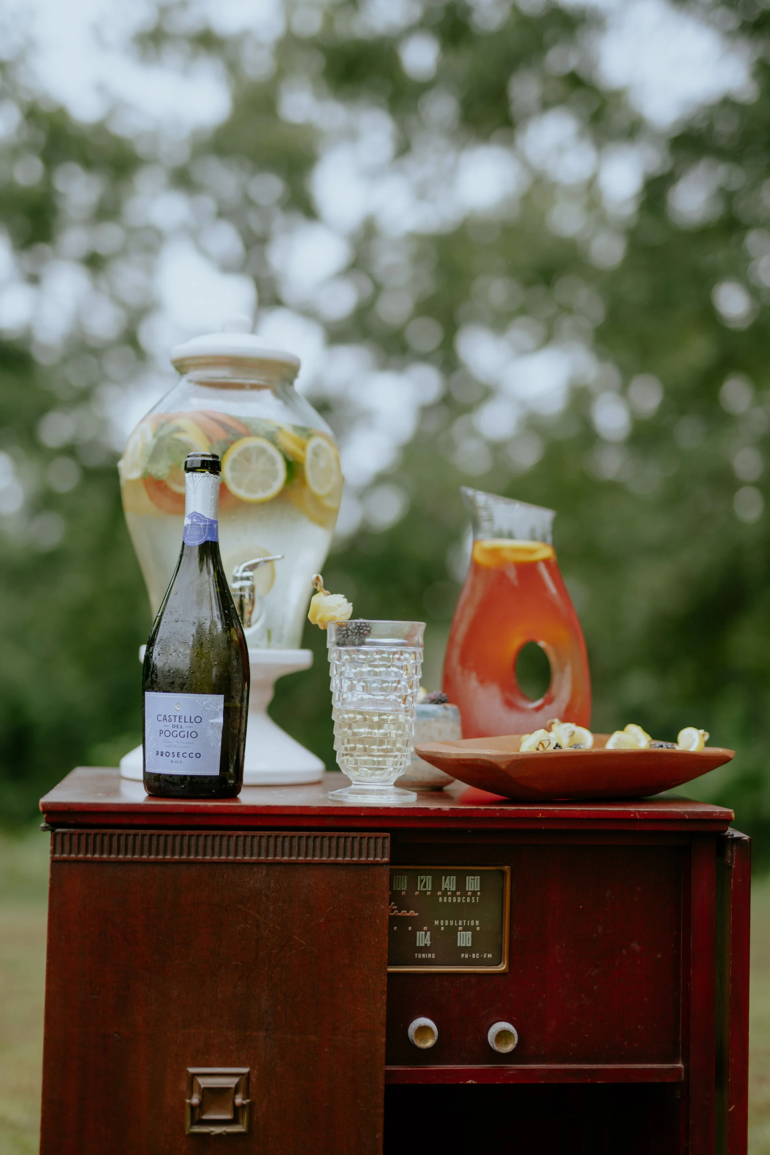 A vintage wooden radio table with a glass pitcher of infused water, a bottle of Prosecco, a glass, and a terracotta-colored decorative vase outside surrounded by green trees.