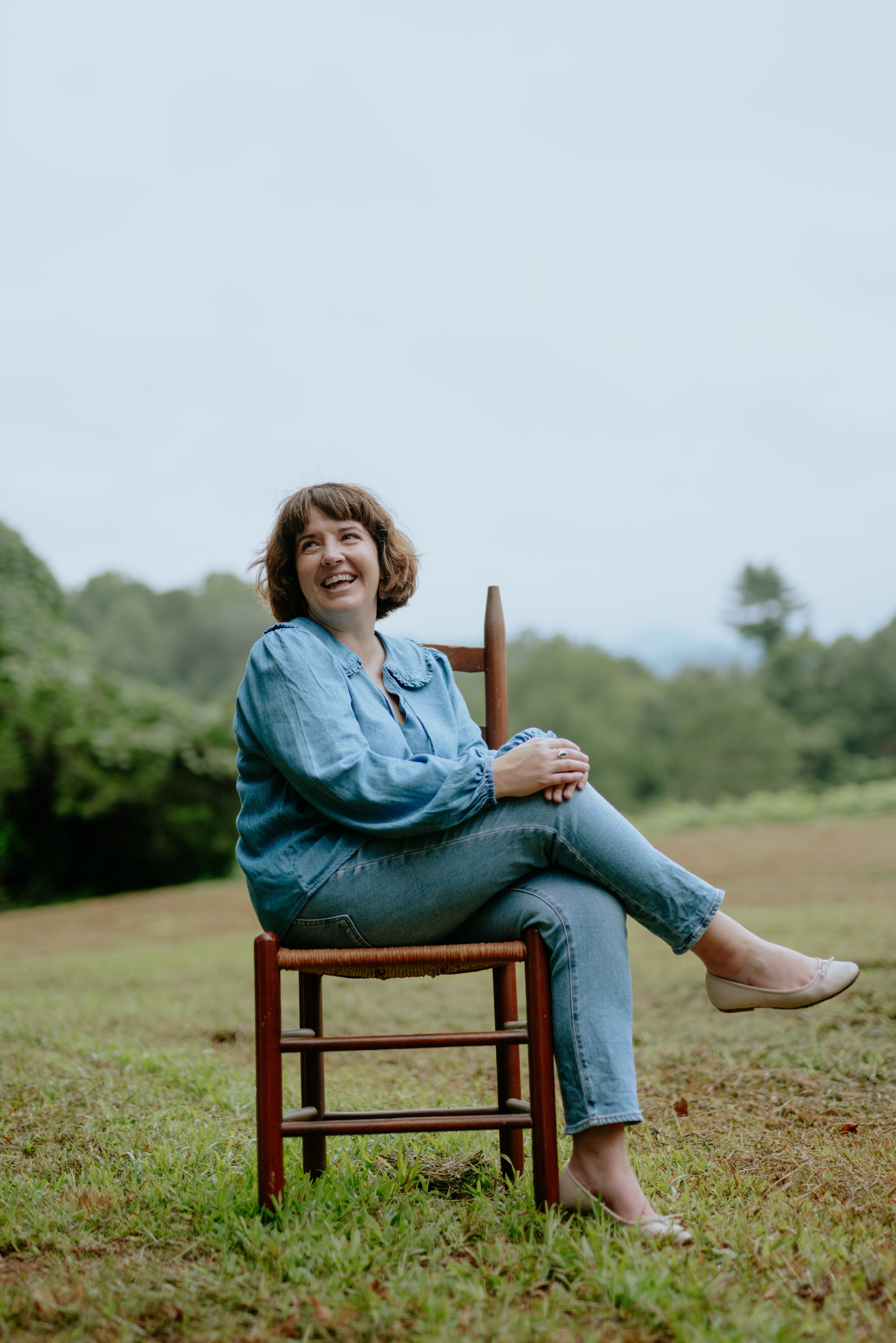 A woman sitting on a wooden chair outdoors on a grassy field, smiling and looking to the side with trees in the background.