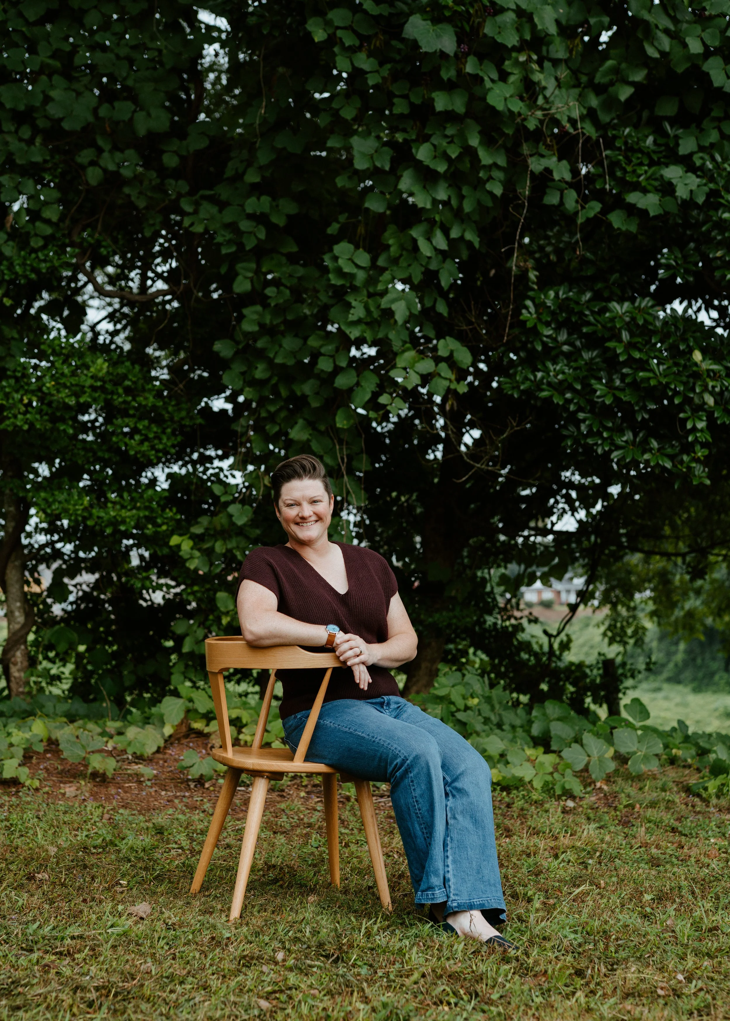 A woman sitting on a wooden chair in a grassy outdoor area, smiling, with a large leafy tree in the background.