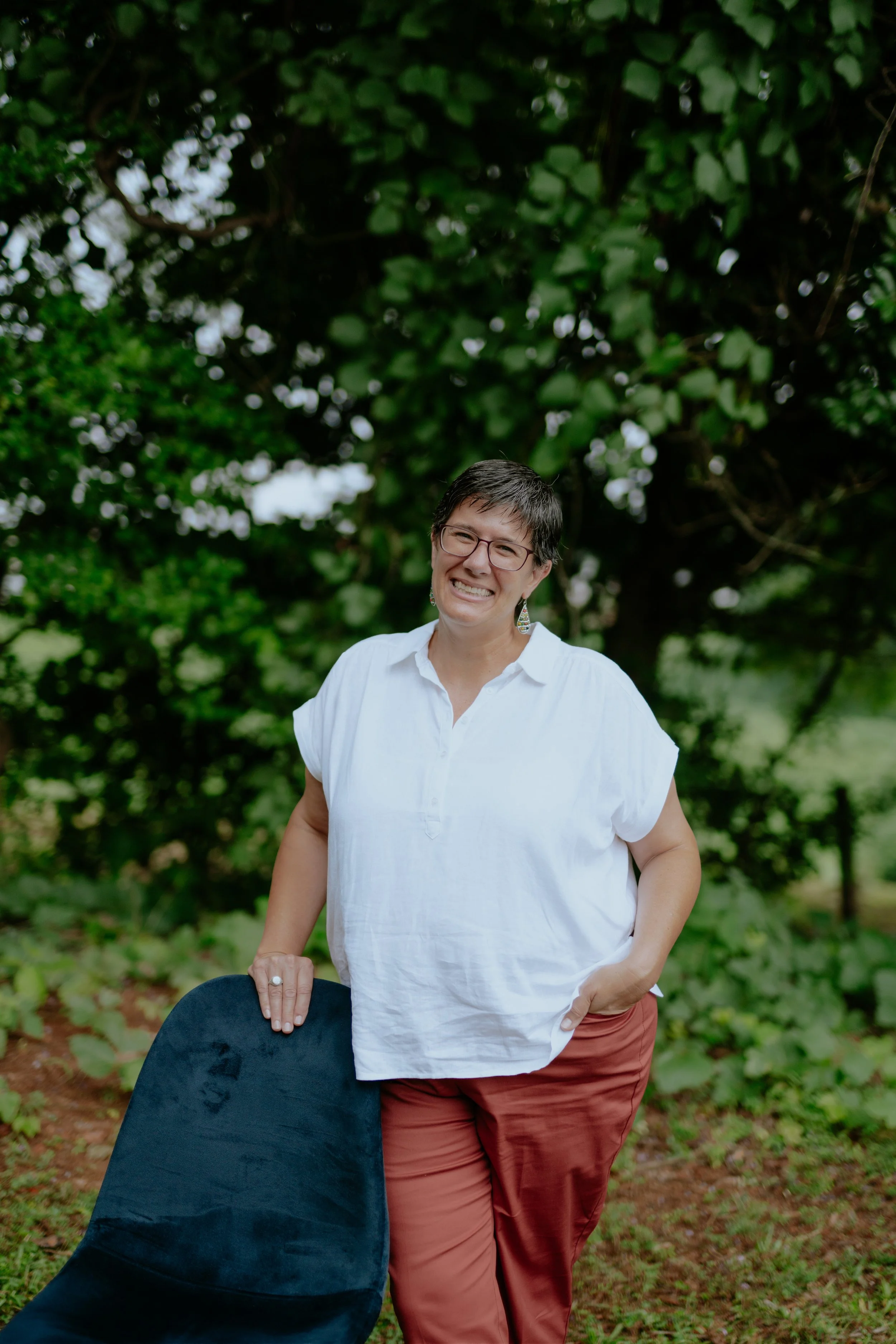 A woman with short dark hair, wearing glasses, a white blouse, and rust-colored pants, smiling outdoors with greenery and trees in the background. She is resting her hand on a black chair.