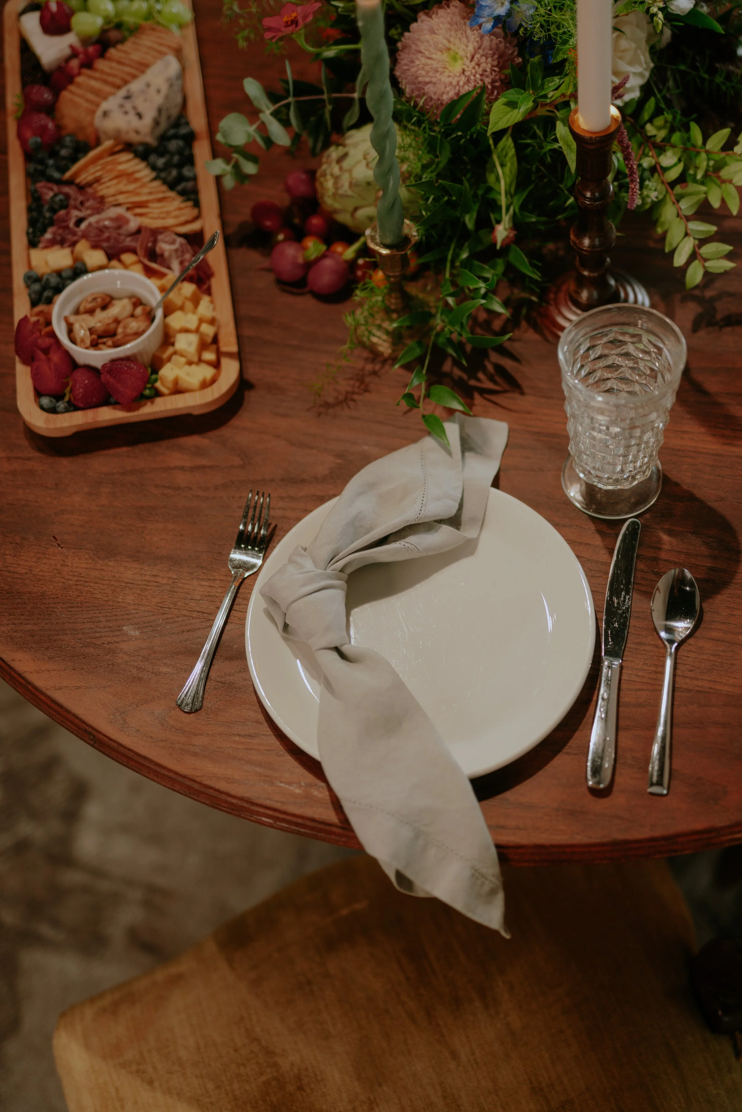 A dining table setting with a white plate, a gray napkin, silver cutlery, a textured glass, and a floral centerpiece, with a charcuterie board featuring cheese, berries, and assorted meats.