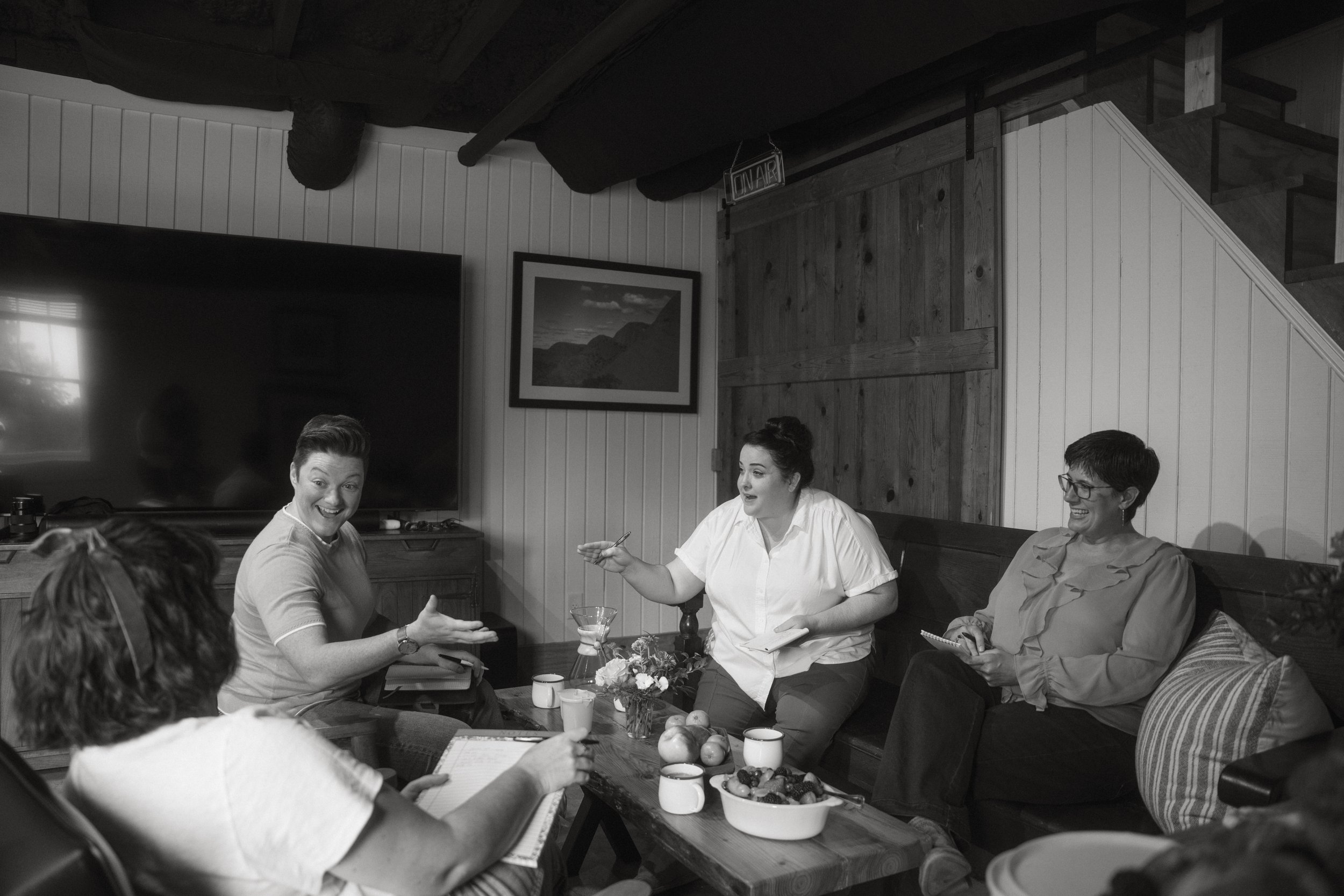 Group of five women sitting around a coffee table, engaged in conversation in a cozy room with wooden walls, one woman animatedly gesturing, and a large flat-screen TV on the wall.