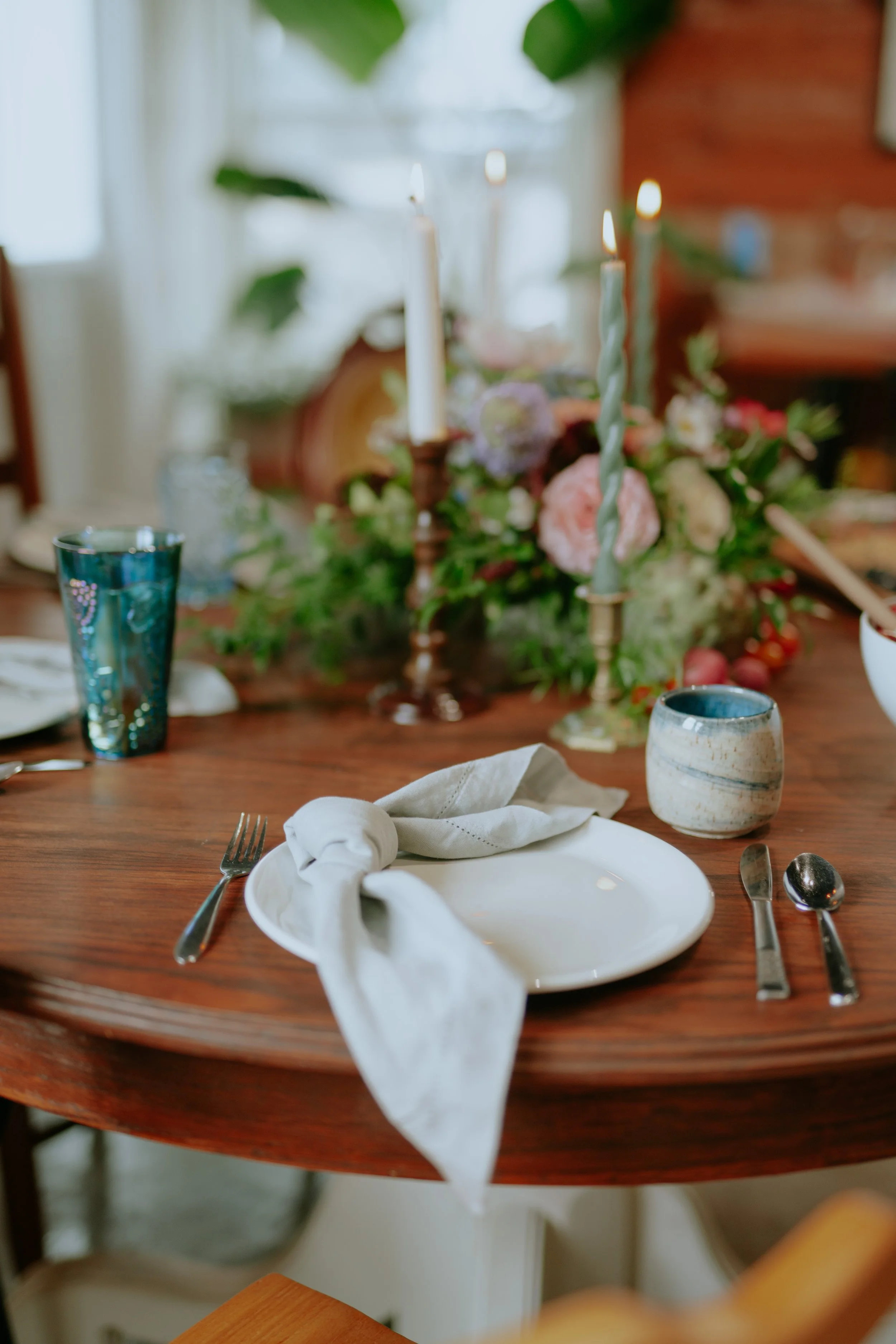 A dining table set with a white plate, a white cloth napkin, a fork, knife, and spoon, with two drinking glasses and a ceramic cup, decorated with a floral centerpiece and lit candles in the background.