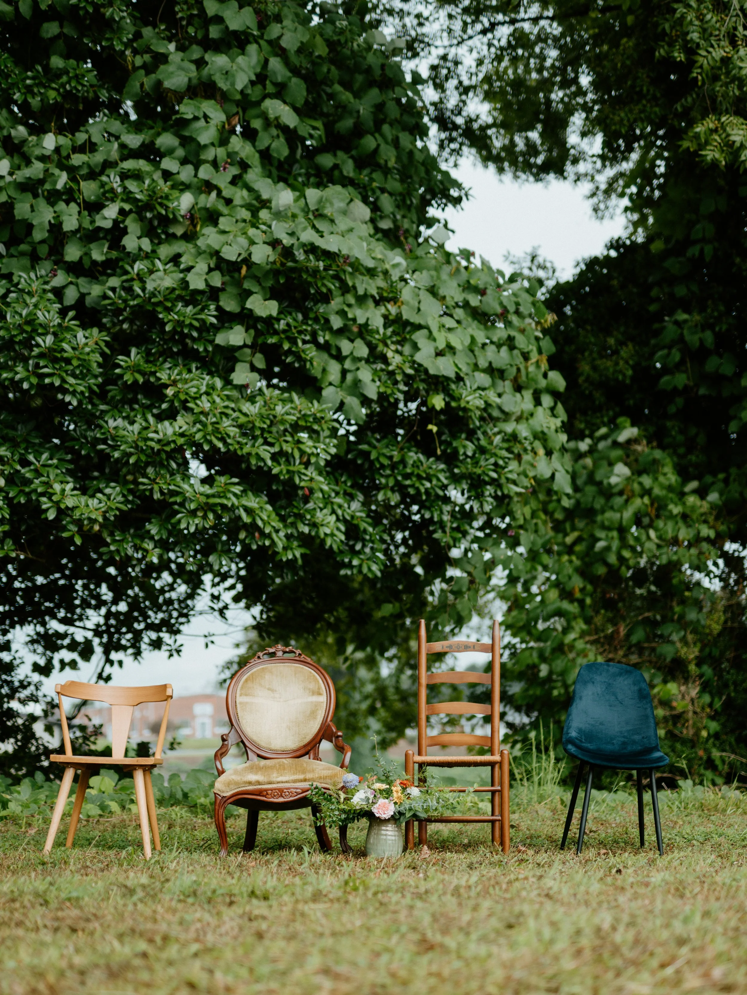 Four chairs of different styles and a bouquet of flowers in a vase, placed outdoors on grass with large green trees in the background.