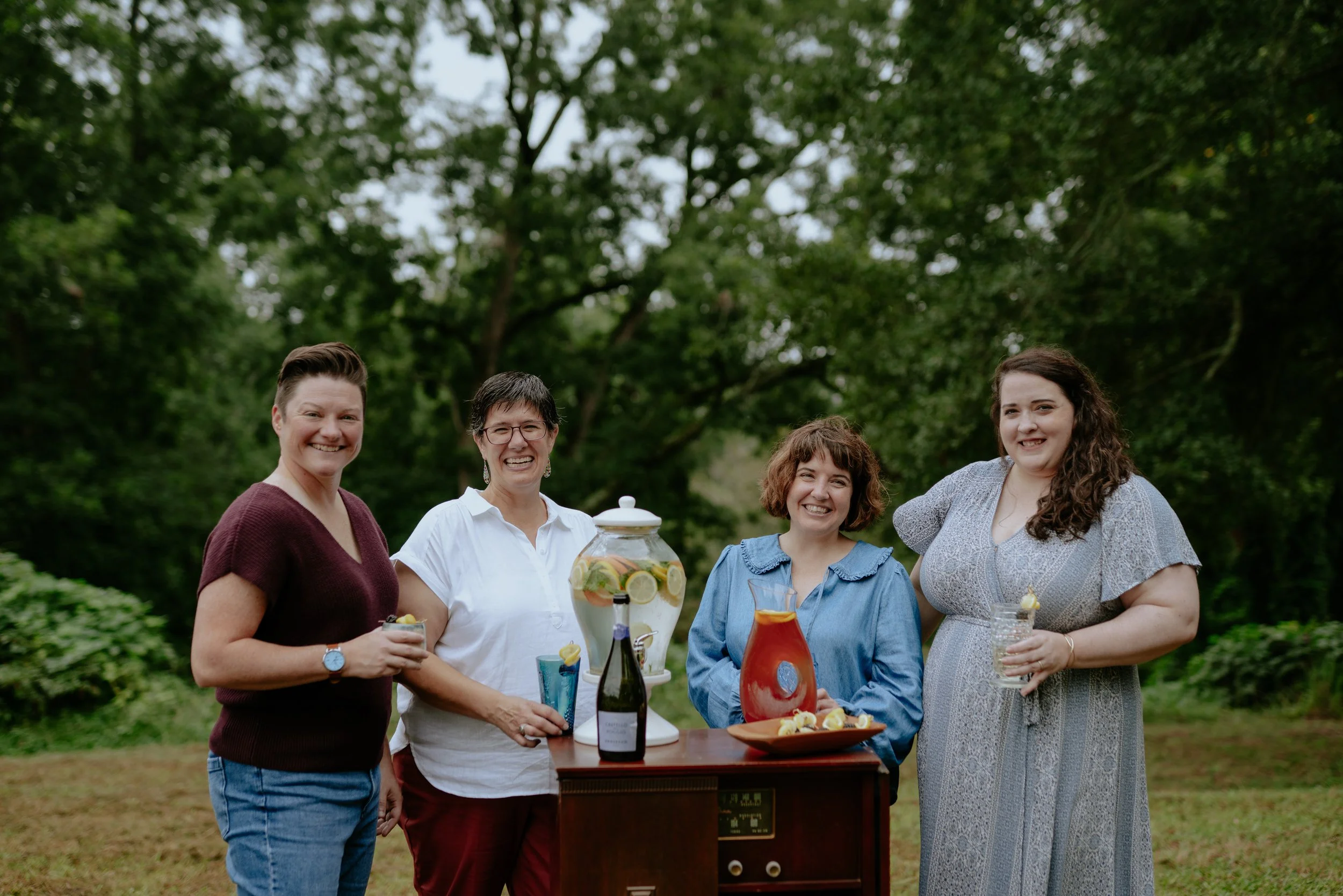 Four women standing outdoors around a small table with drinks and a beverage dispenser, smiling at the camera.