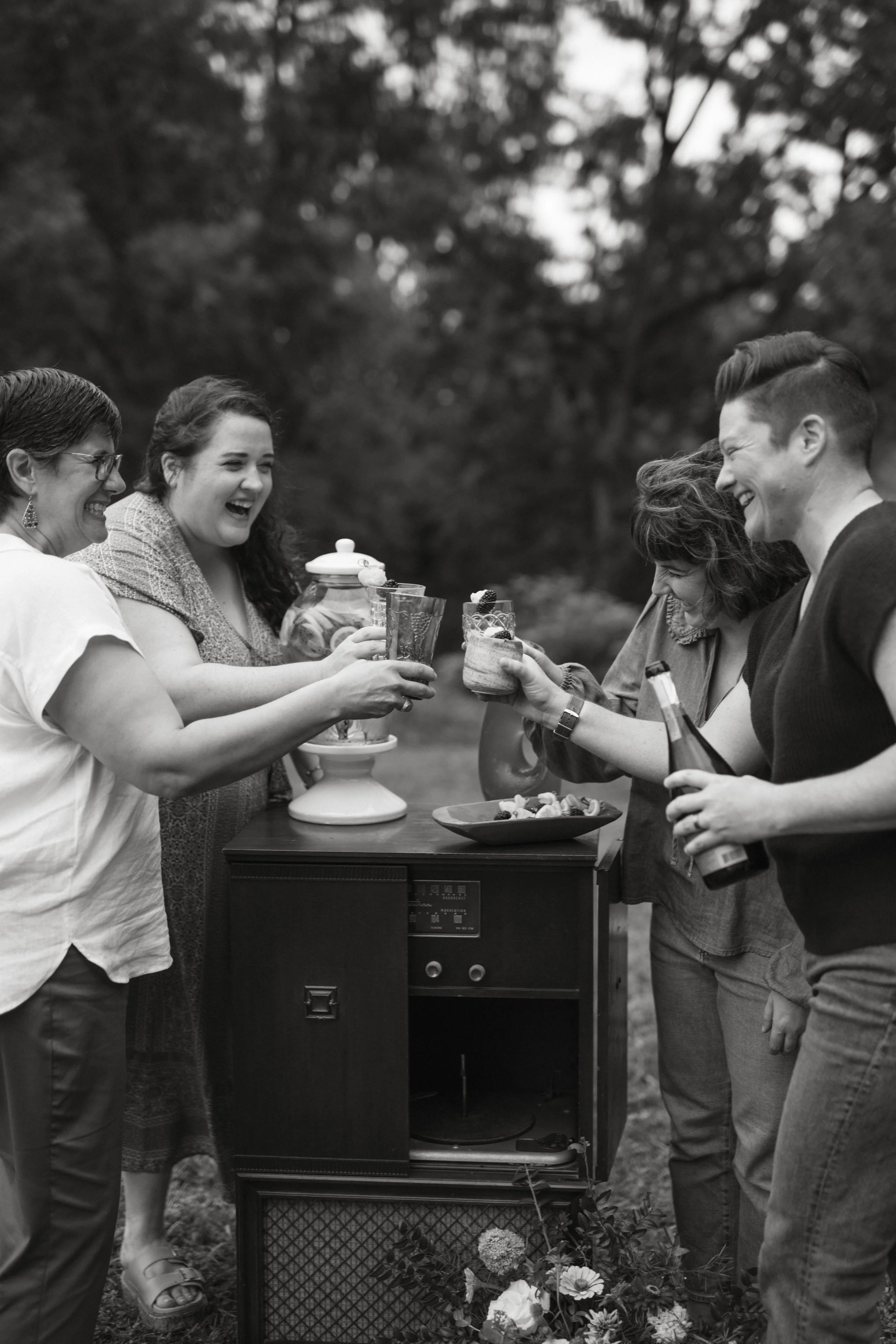Group of five friends enjoying drinks and snacks around a vintage record player outdoors, smiling and toasting, in a park setting.