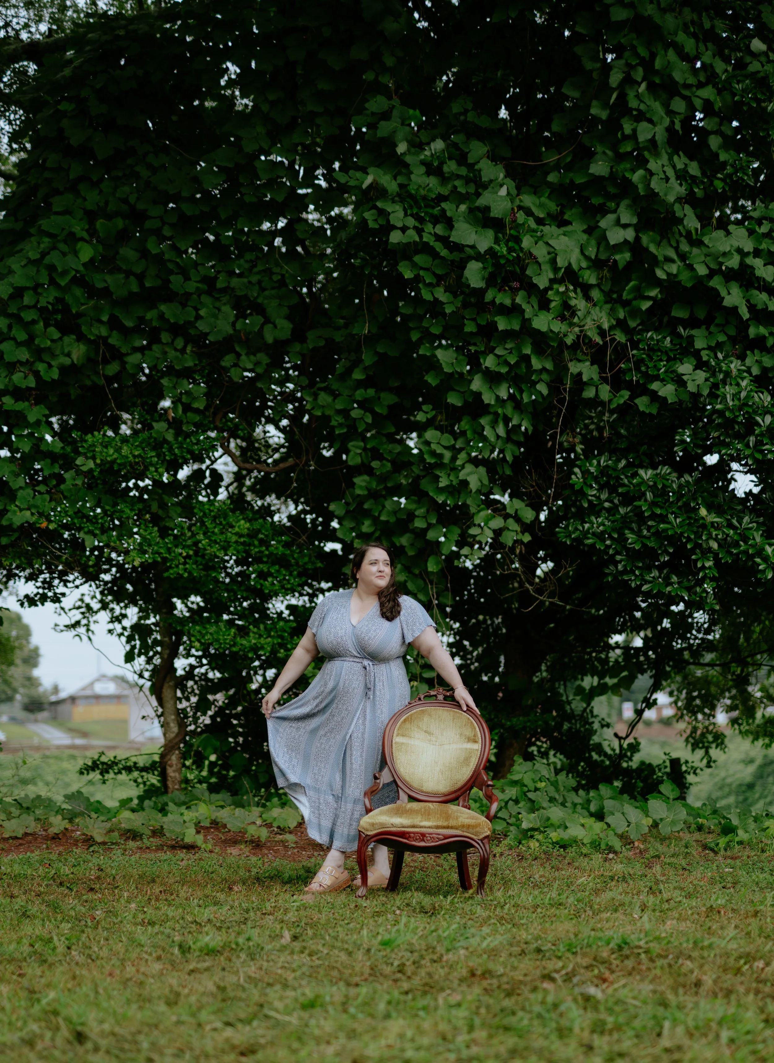 A woman in a long, patterned dress standing on grass beside an antique armchair, with a large leafy tree in the background.