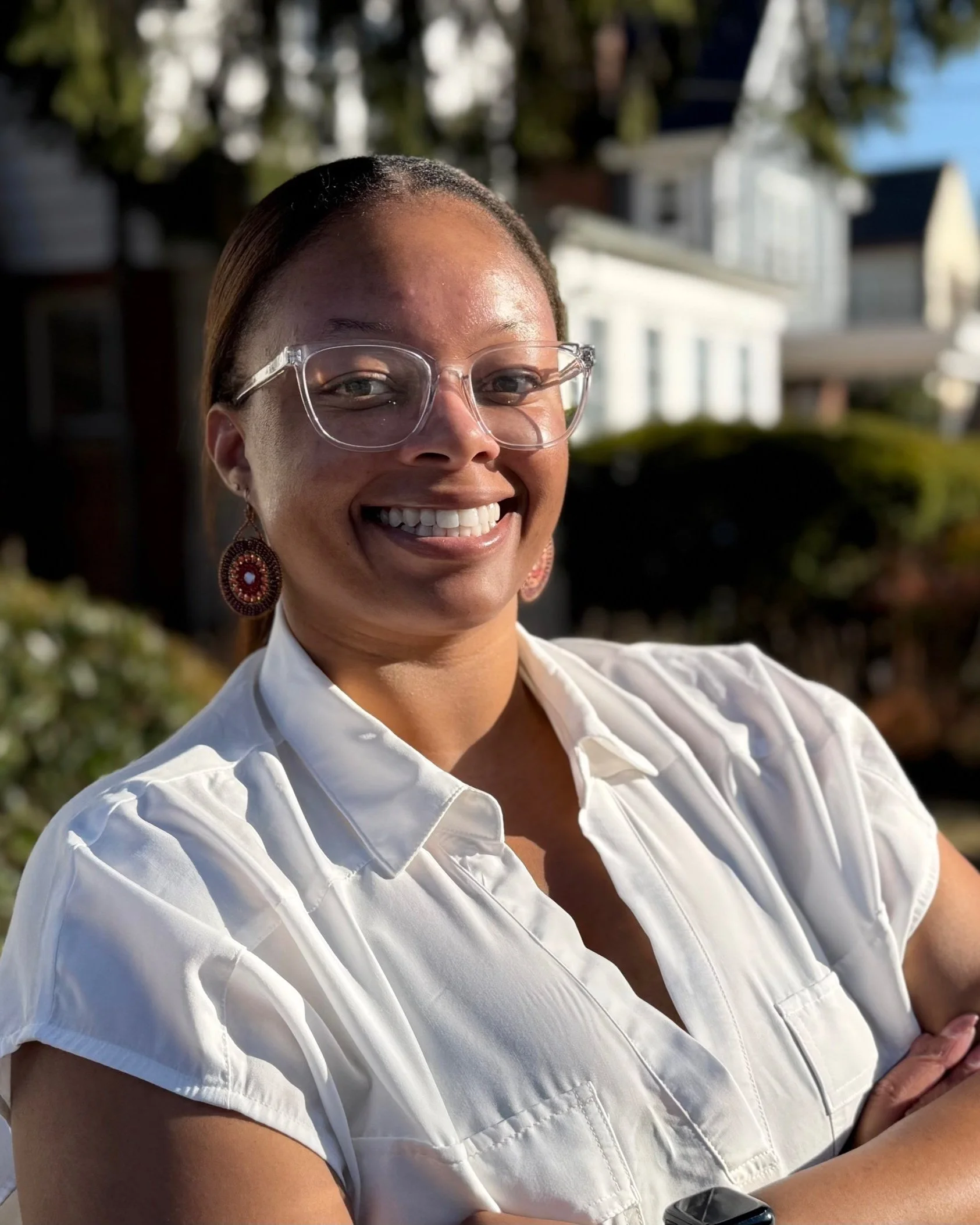 A smiling woman wearing glasses, earrings, and a white blouse outdoors with houses and trees in the background.