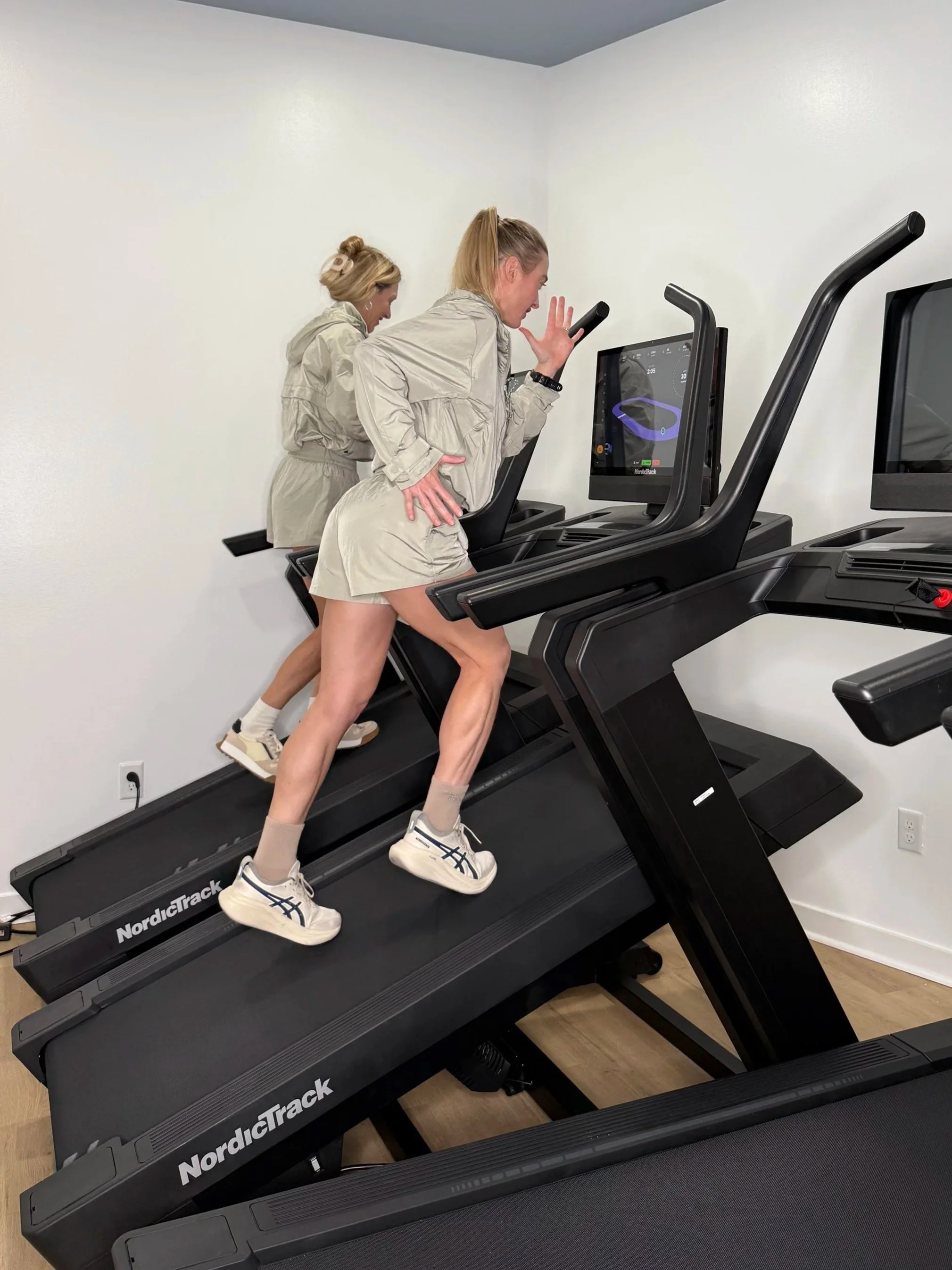 Two women running on NordicTrack treadmills in a room with white walls and wooden floor. One woman is looking at her phone, and the other is mid-step on the treadmill.