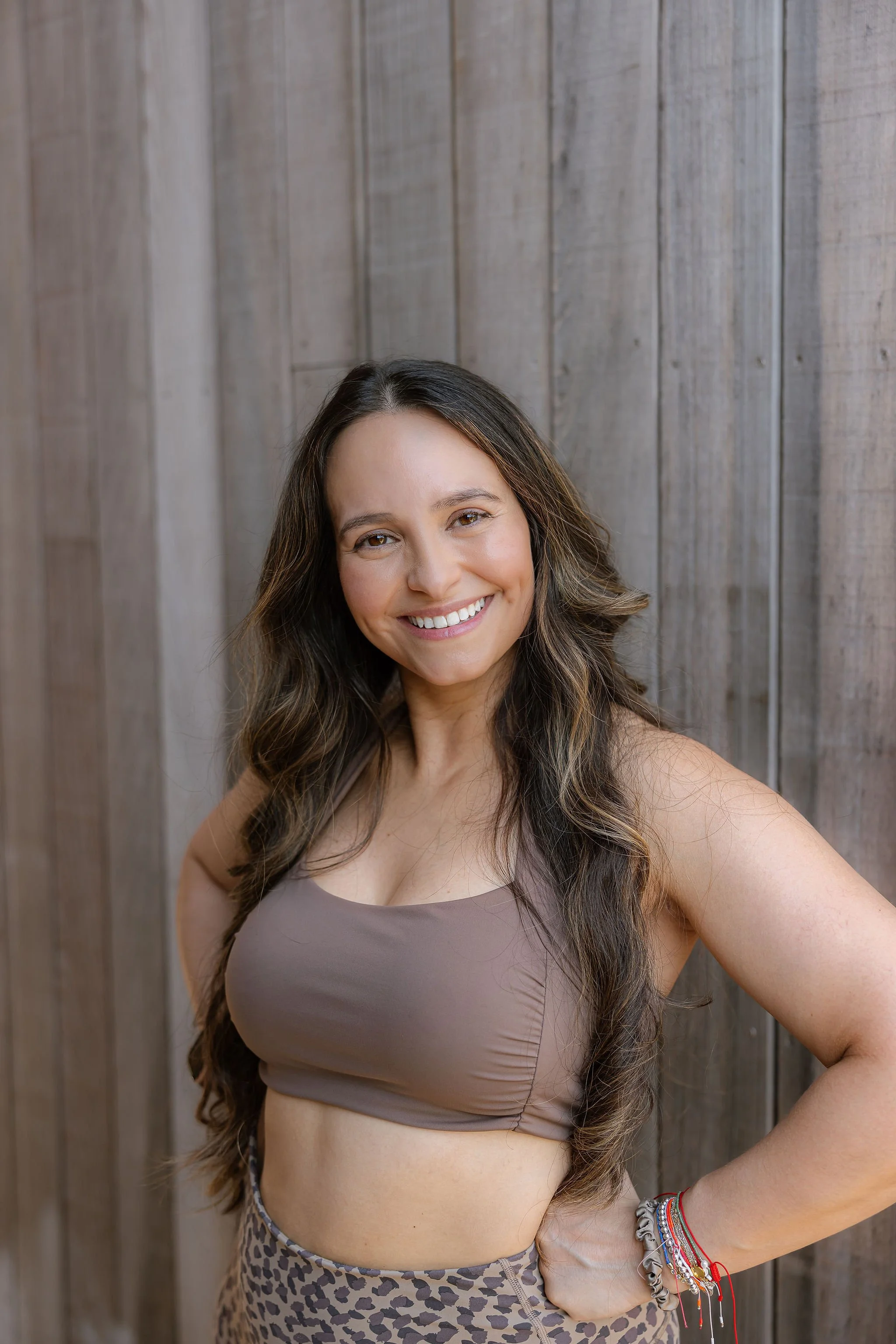 A woman with long wavy hair smiling at the camera, standing in front of a wooden wall, wearing a beige crop top and leopard print pants, accessorized with bracelets.