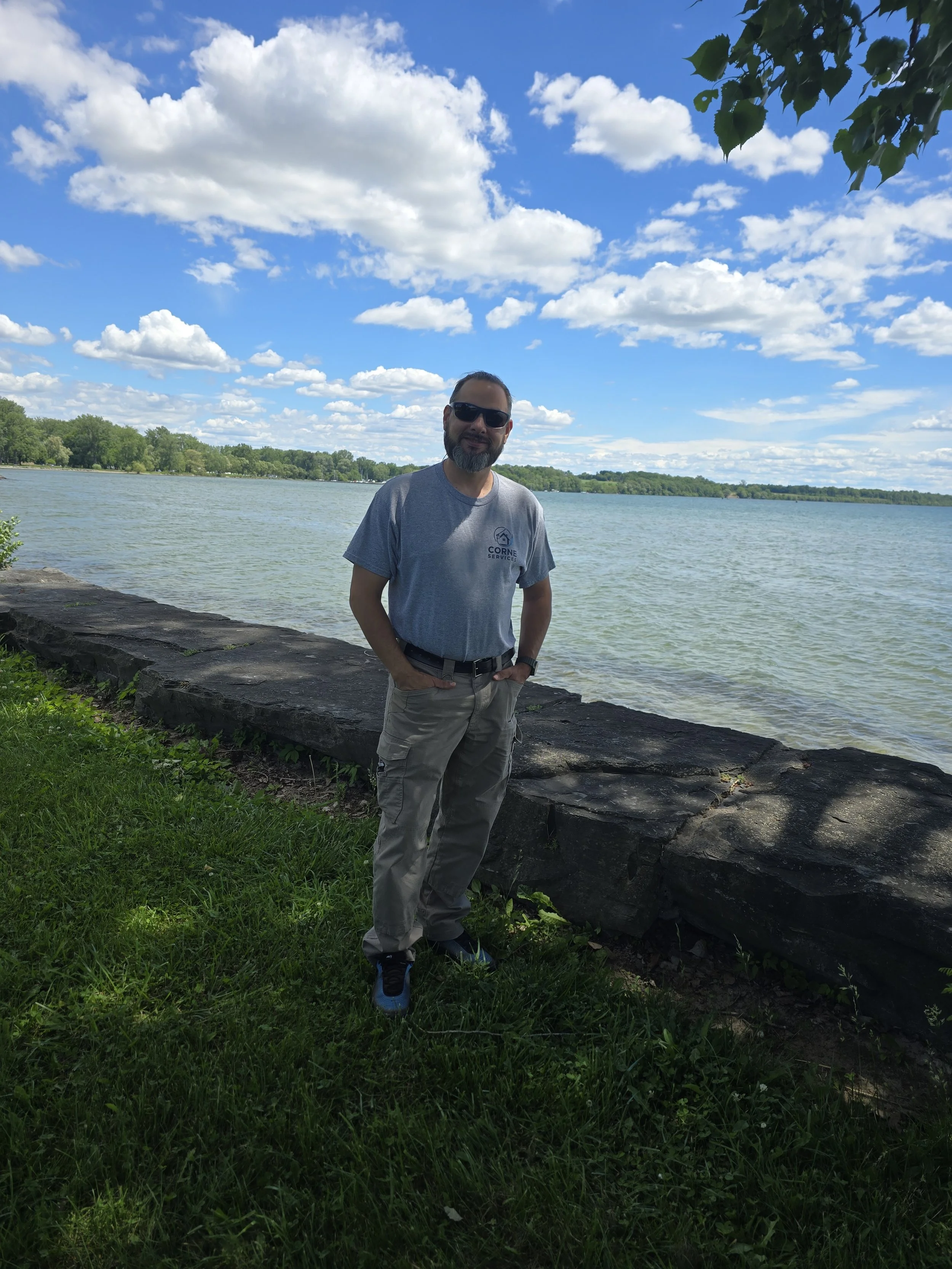 A man wearing sunglasses, a gray T-shirt, and beige cargo pants standing by a large body of water with trees in the background under a partly cloudy sky.