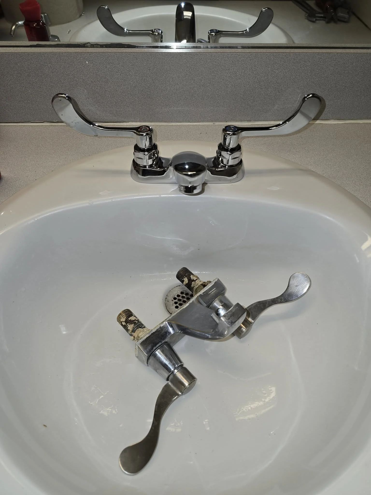 A bathroom sink with a chrome faucet and a damaged faucet lying inside the sink basin.