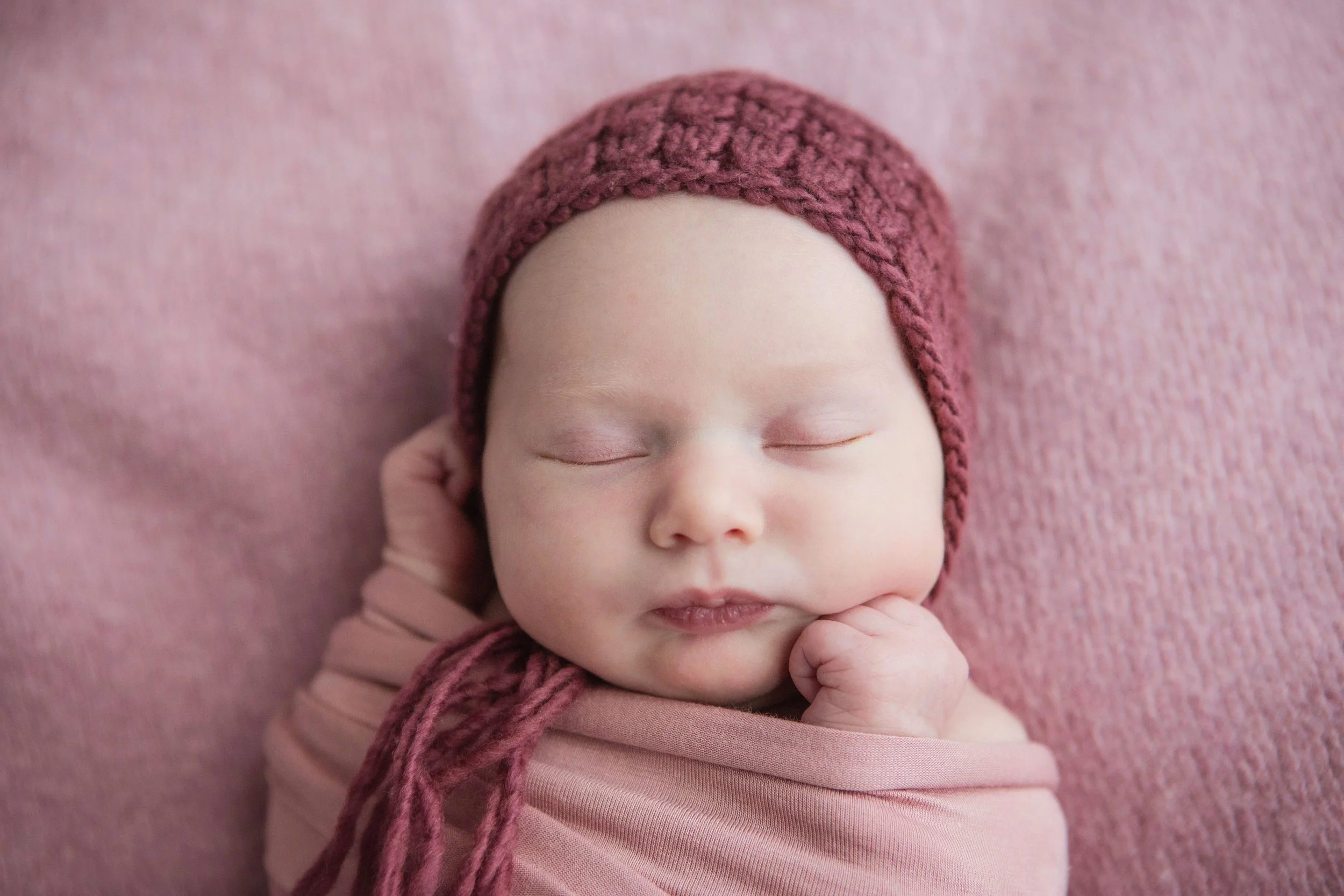 A close-up of a sleeping newborn baby with a knitted pink hat and wrapped in pink cloth, resting peacefully on a pink textured background.