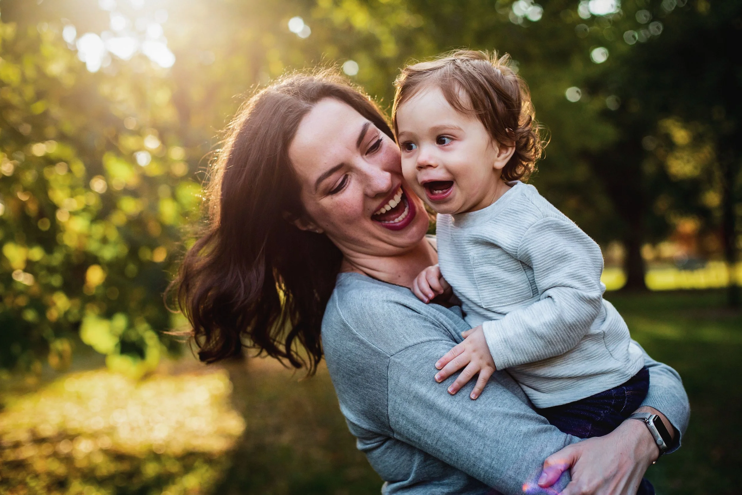A woman with brown hair and a woman with curly brown hair in a gray shirt smiling and playing with a young boy in a gray sweatshirt outdoors with trees and sunlight in the background.