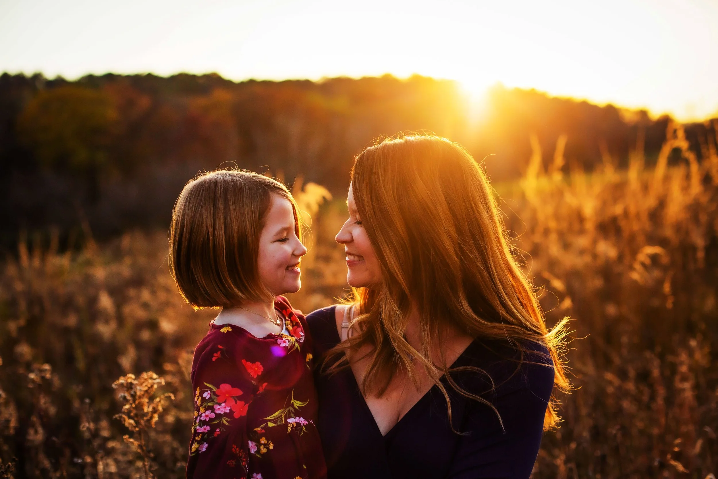 A woman and a young girl face each other smiling in a field at sunset, with warm sunlight in the background.