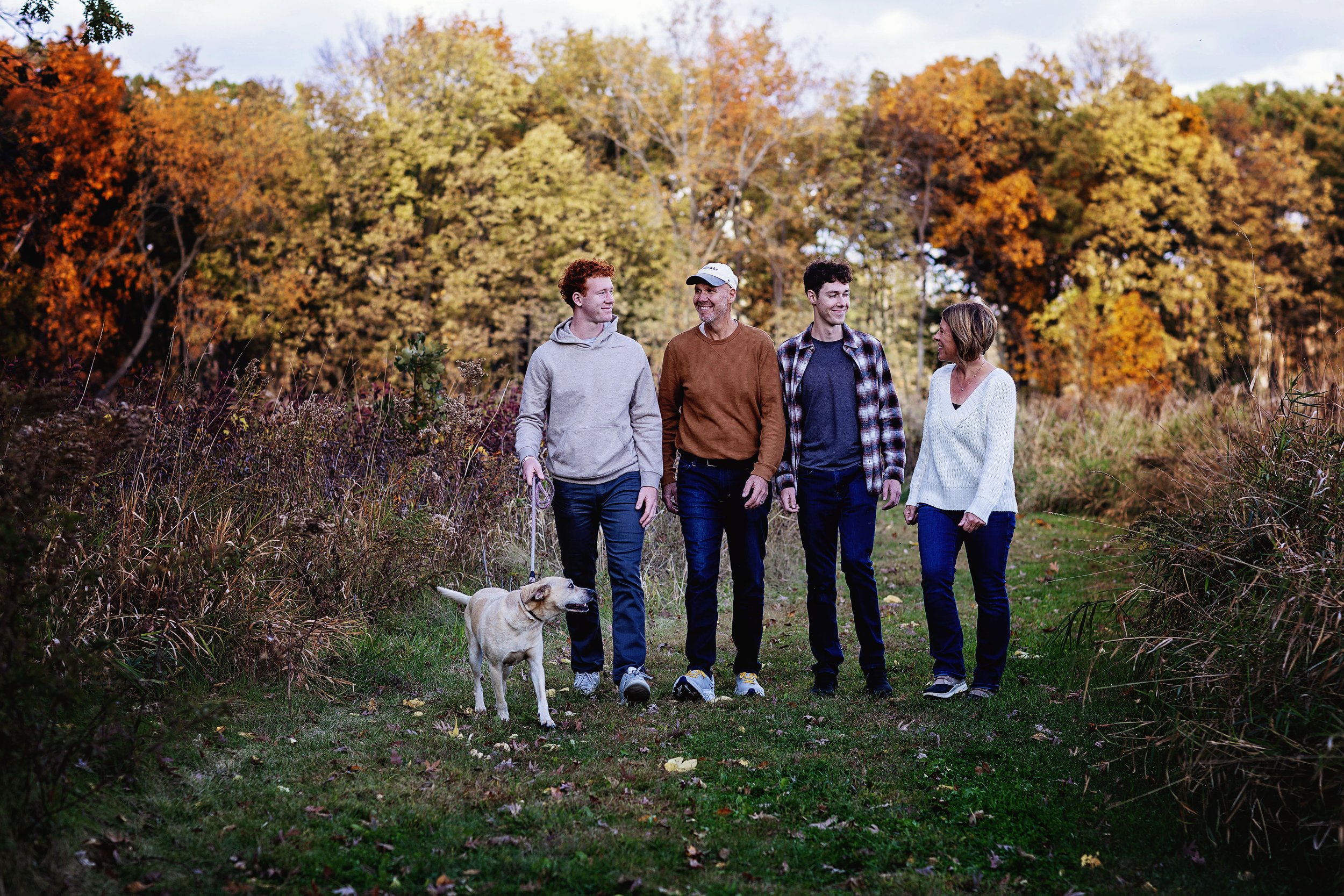 Four people walking outdoors during autumn with a dog, surrounded by colorful fall trees and foliage.