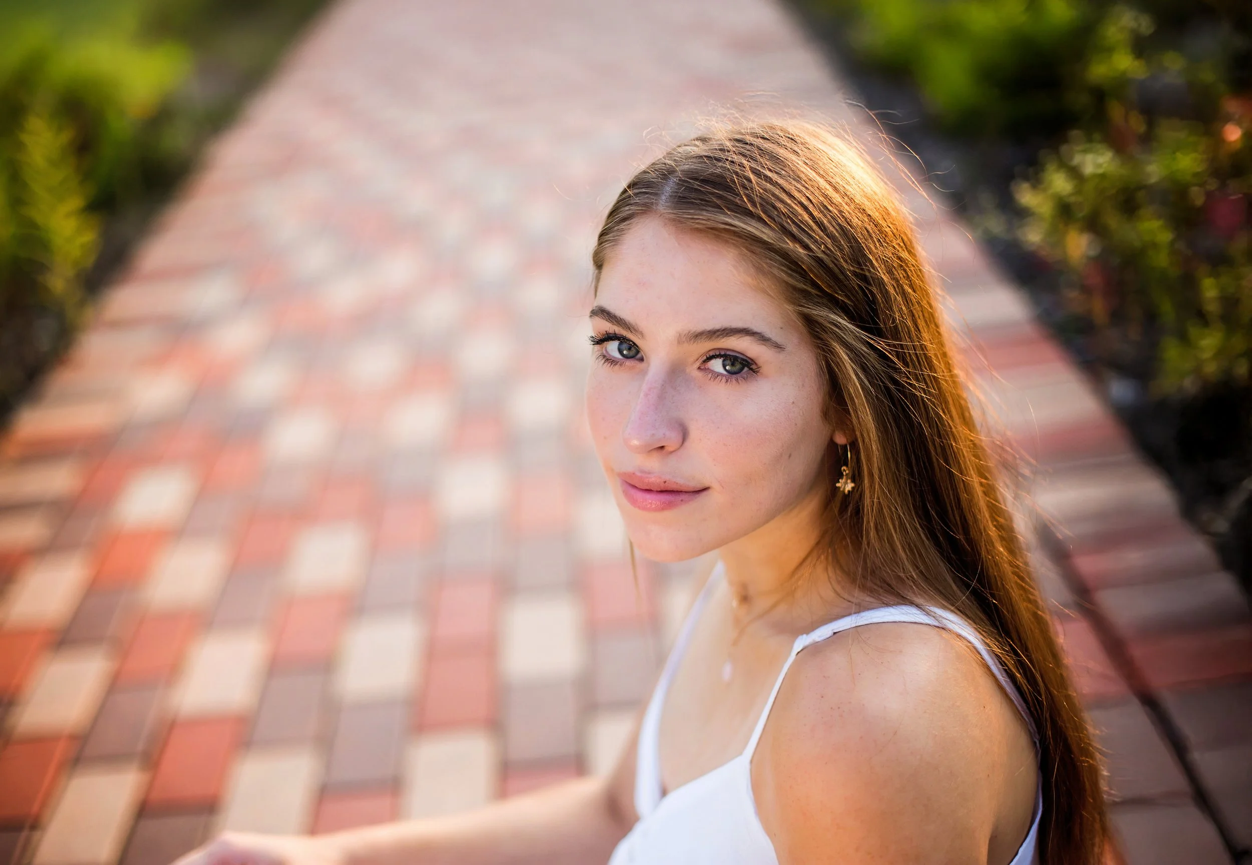 A young woman with long red hair and blue eyes looking up at the camera, standing outdoors on a brick pathway with greenery on the sides.
