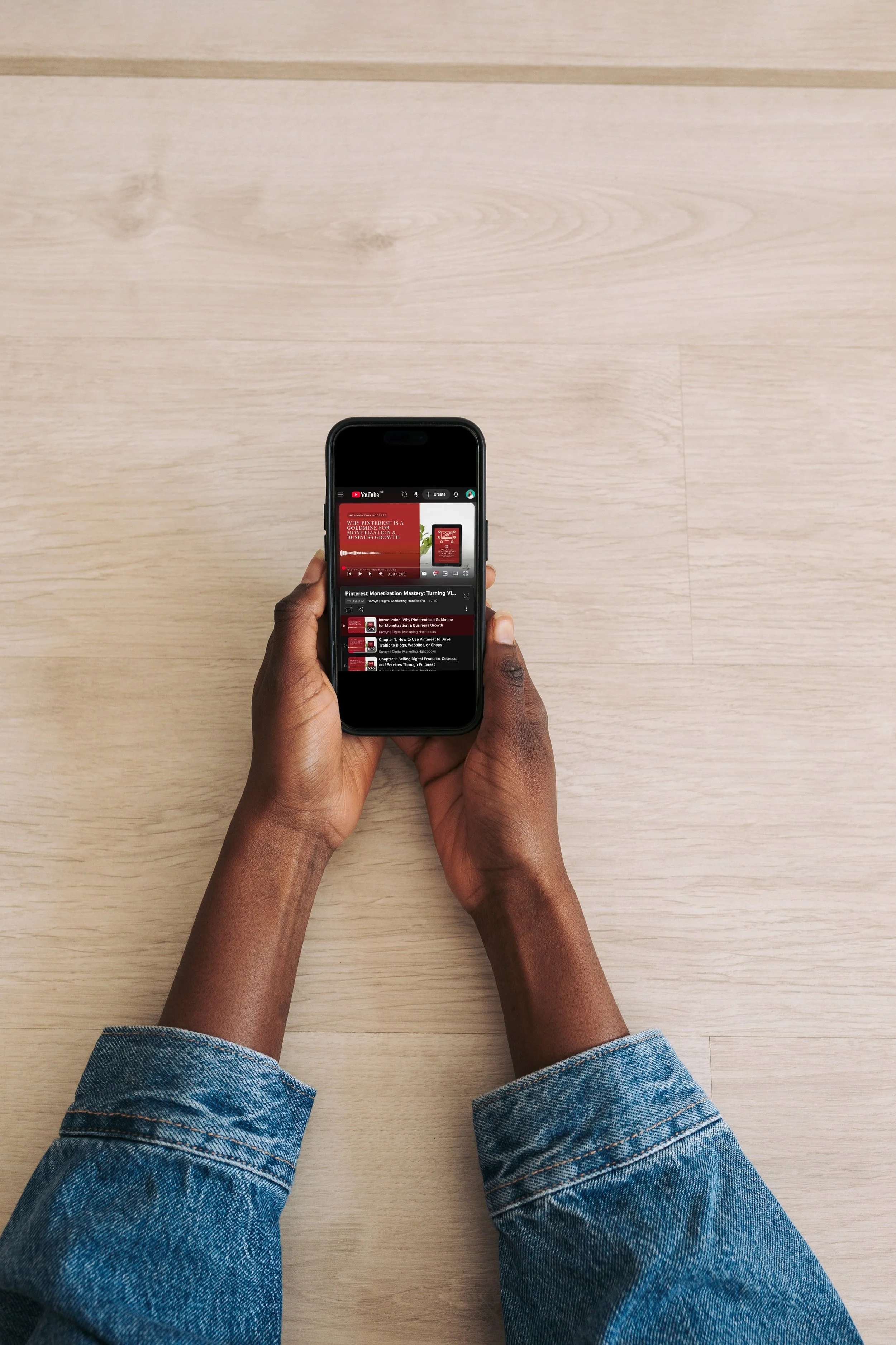 Person holding a smartphone, browsing YouTube, with a light wooden table in the background.
