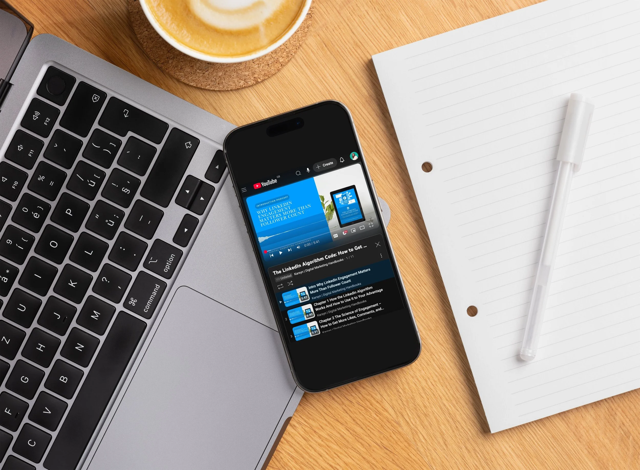 Laptop keyboard, smartphone playing a YouTube video, notepad with a pen, and a cup of coffee on a wooden desk.