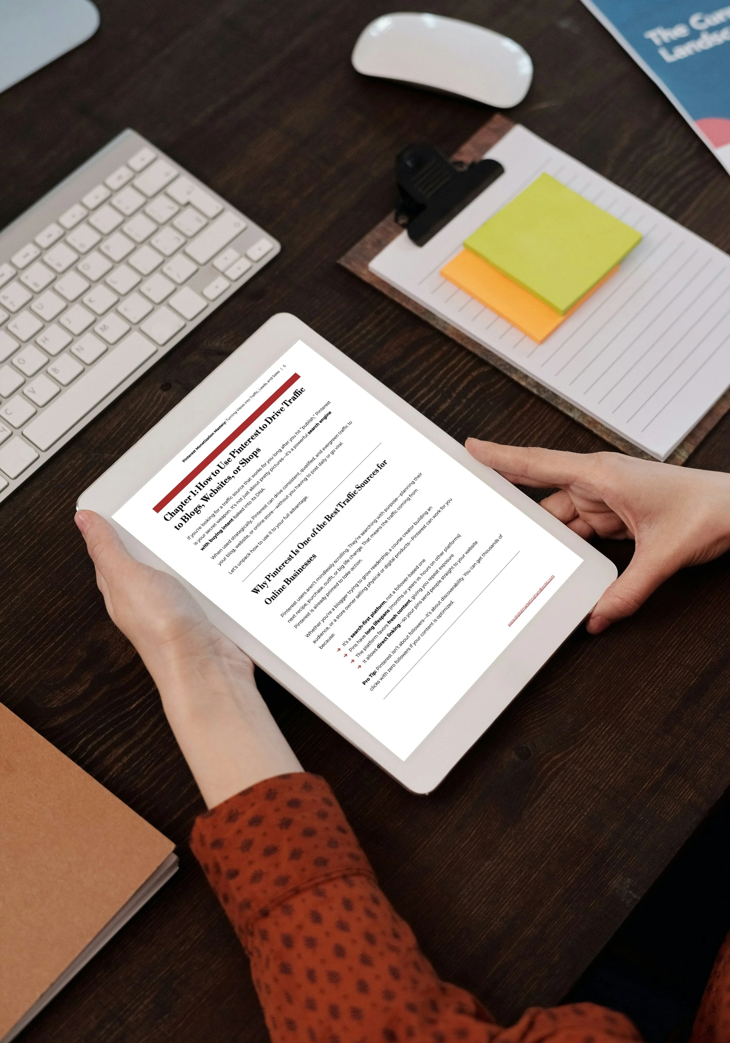 Person reading an article on a tablet at a wooden desk, surrounded by a white keyboard, a white mouse, sticky notes, a clip, and a notebook.