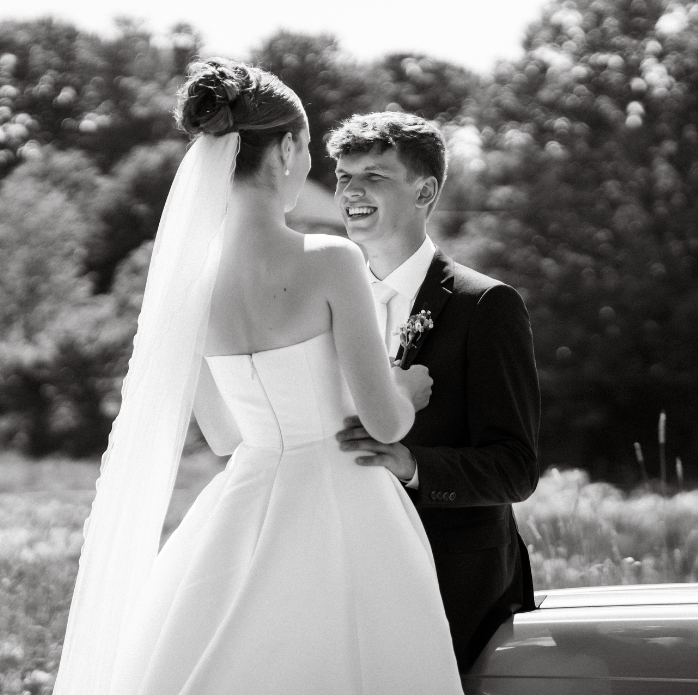 A bride in a strapless wedding dress with a veil and a groom in a black tuxedo smiling at each other outdoors.