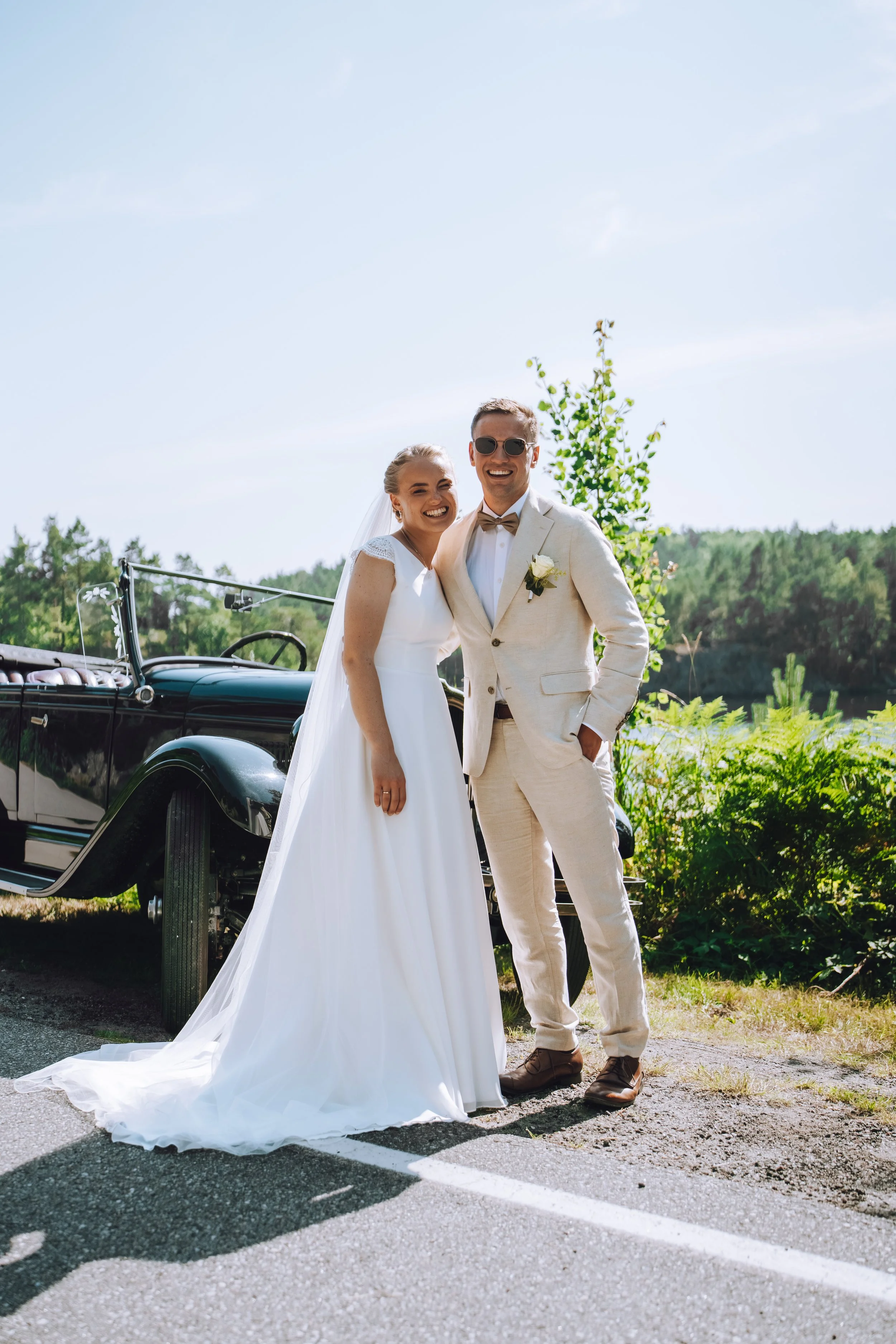 Et brudepar smiler foran en veteranbil med grønne trær i bakgrunnen, en solrik dag, på en vei med hvite linjer.