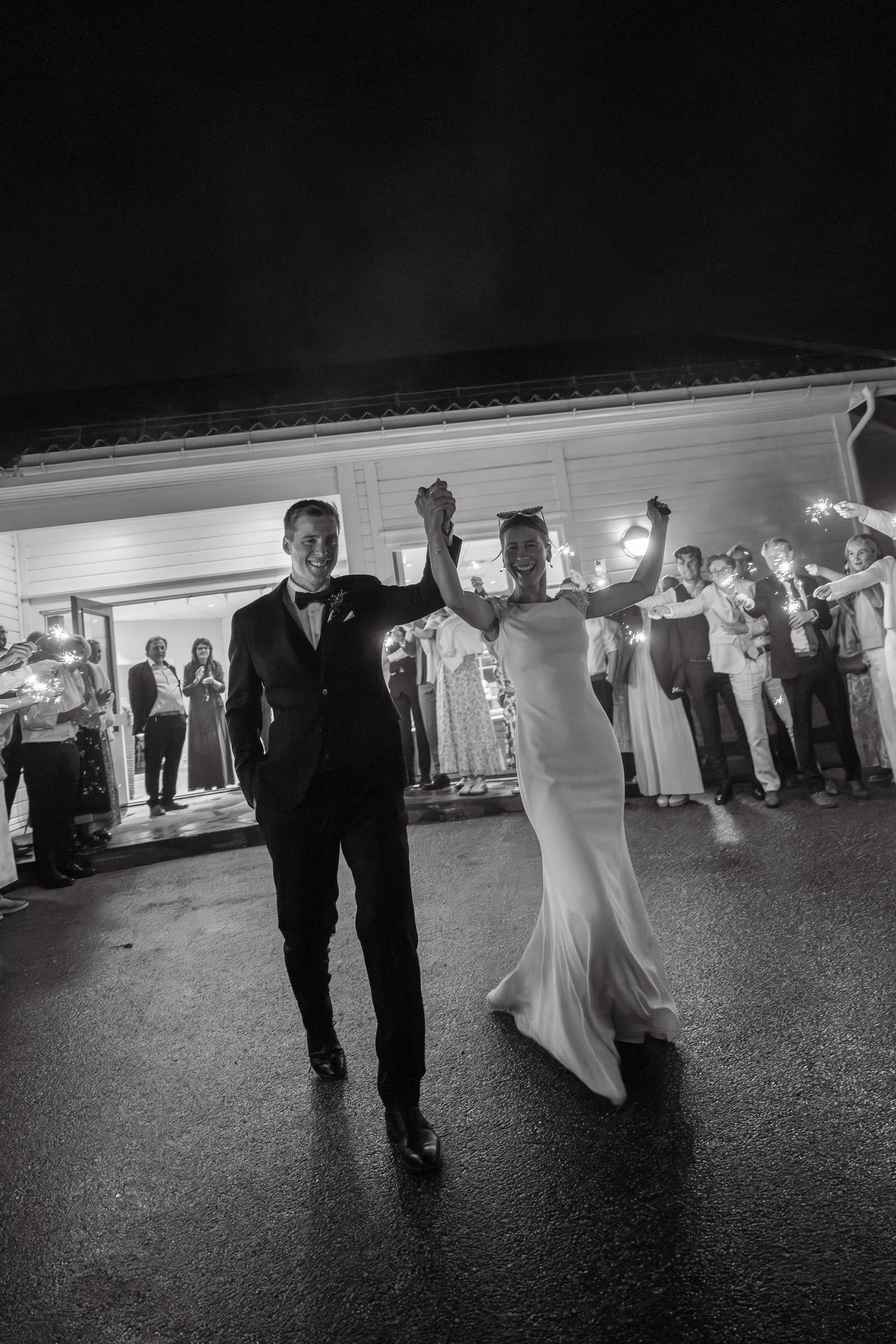 En lykkelig brud og brudgom danser sammen under en bryllupsfeiring, omgitt av familie og venner som holder fakler utendørs om kvelden.