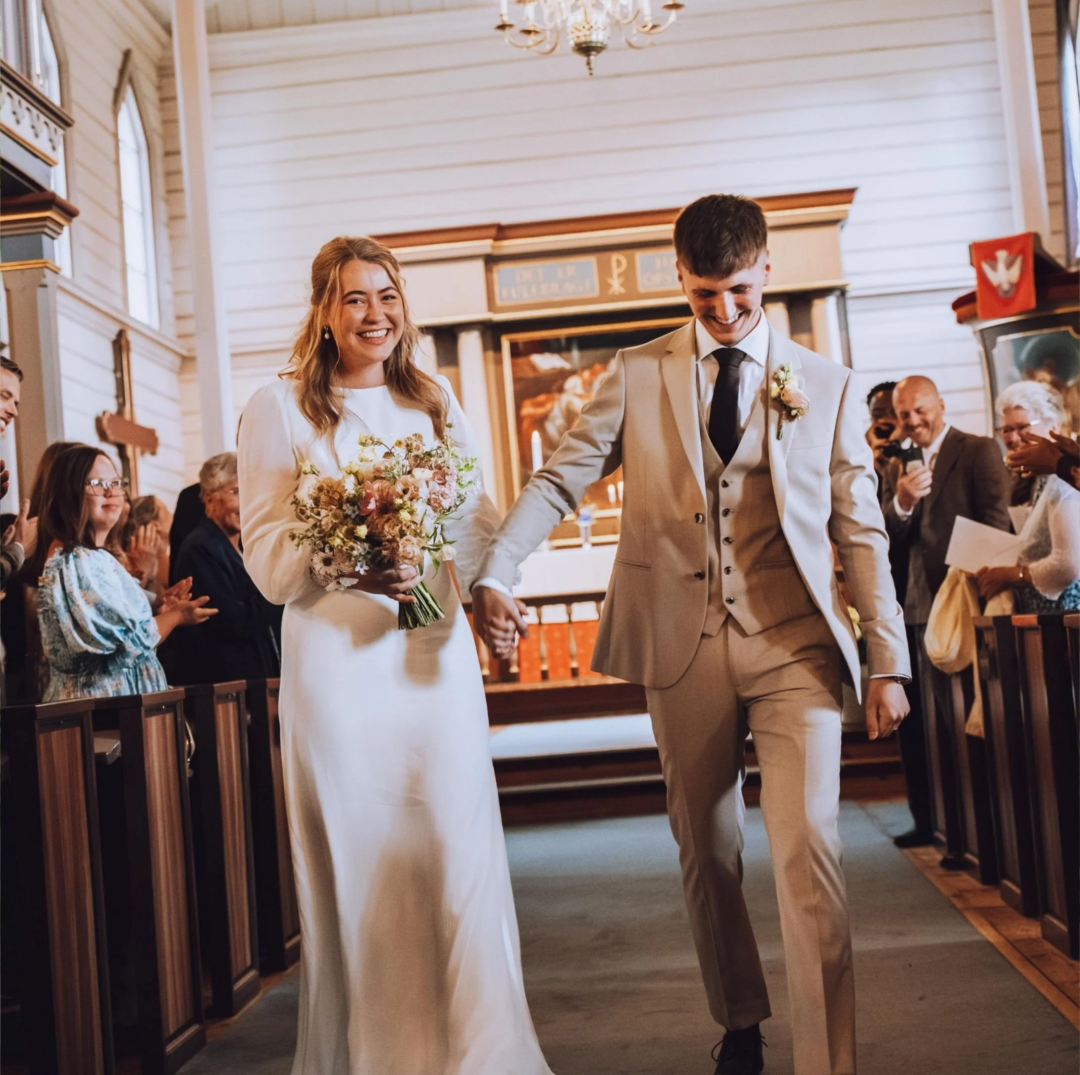 Bride and groom walking down the aisle in a church, holding hands, smiling, surrounded by seated guests clapping and smiling.