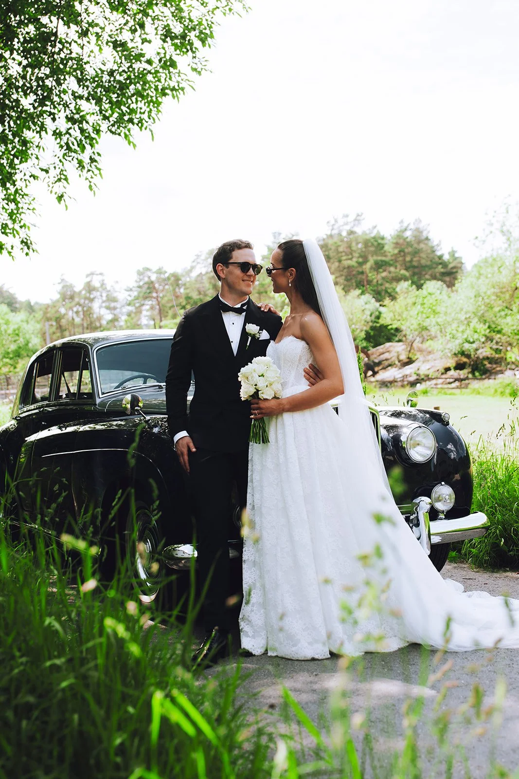 Et brudepar står sammen foran en svart klassisk bil i en grønn park. Bruden har lang hvit kjole og holder en bukett hvite blomster, mens brudgommen har på seg dress og solbriller.